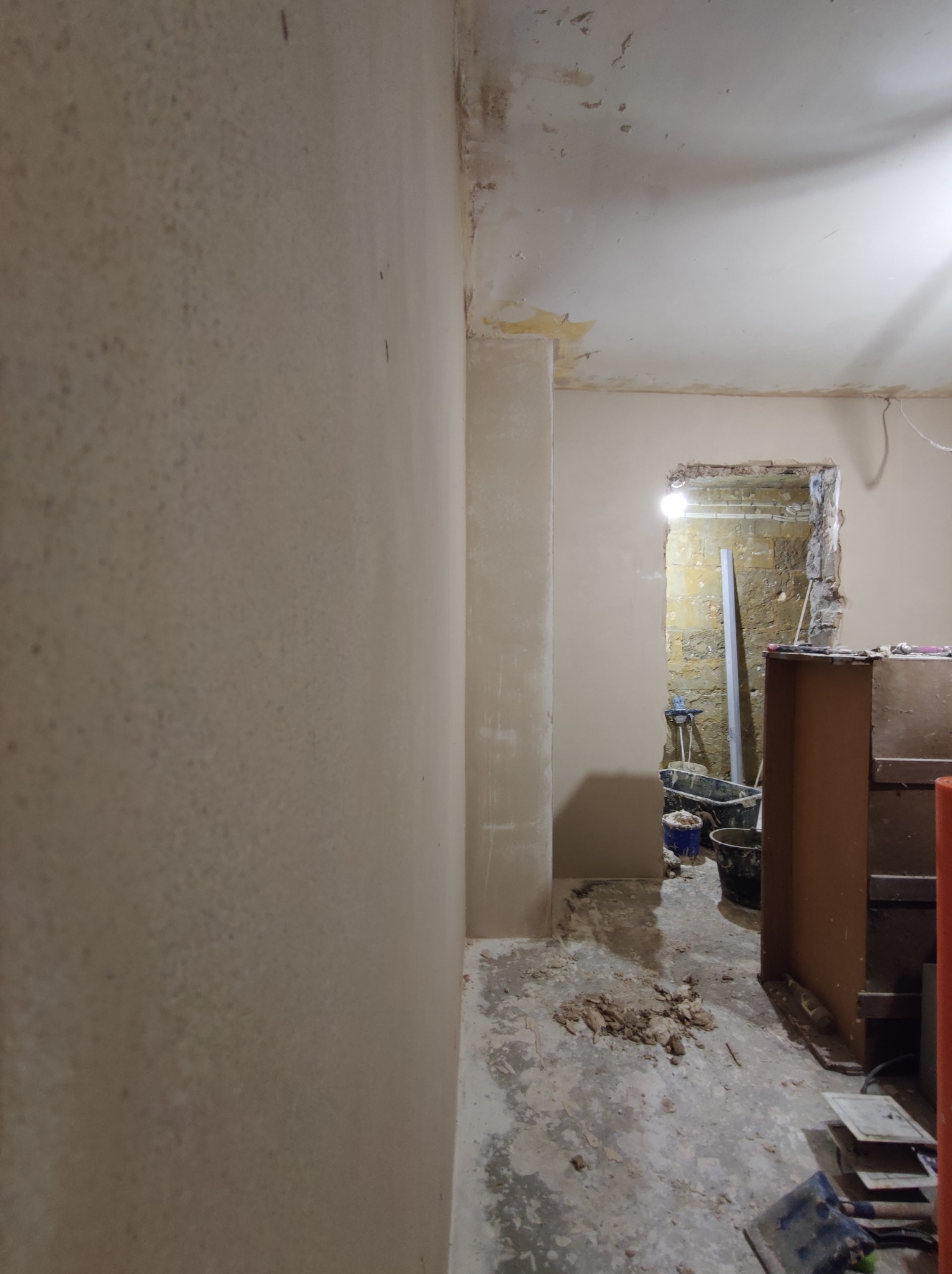 Wnętrze w trakcie remontu: ściany częściowo pokryte gładzią, widoczne nierówności i pozostałości po pracach, na podłodze gruz i narzędzia budowlane, w tle widoczne wiaderka i inne akcesoria.