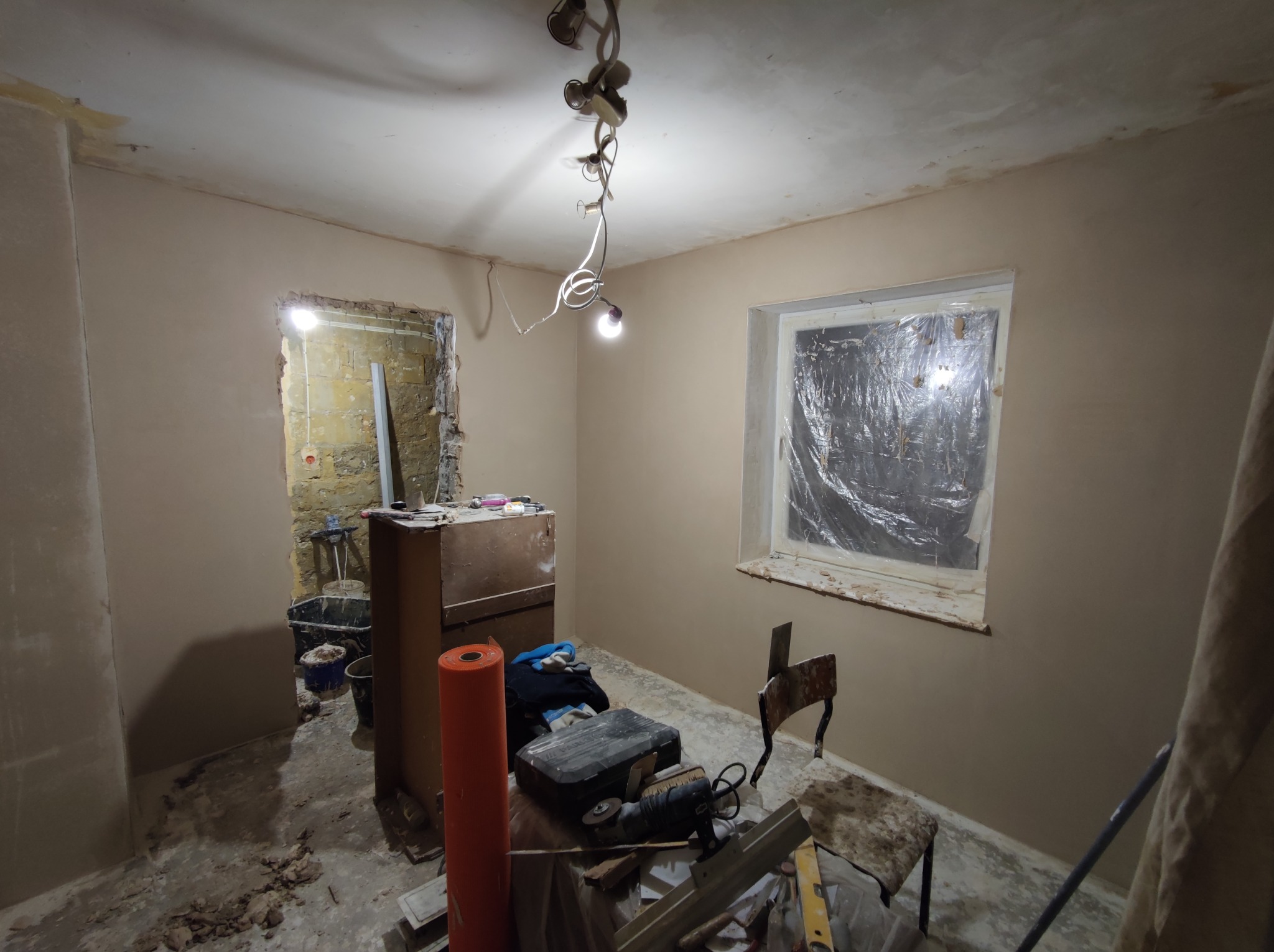 Pokój w trakcie remontu: ściany pokryte świeżo nałożoną gładzią, okno zabezpieczone folią, widoczne narzędzia, wiadro i resztki materiałów na podłodze, surowa żarówka zwisająca z sufitu.