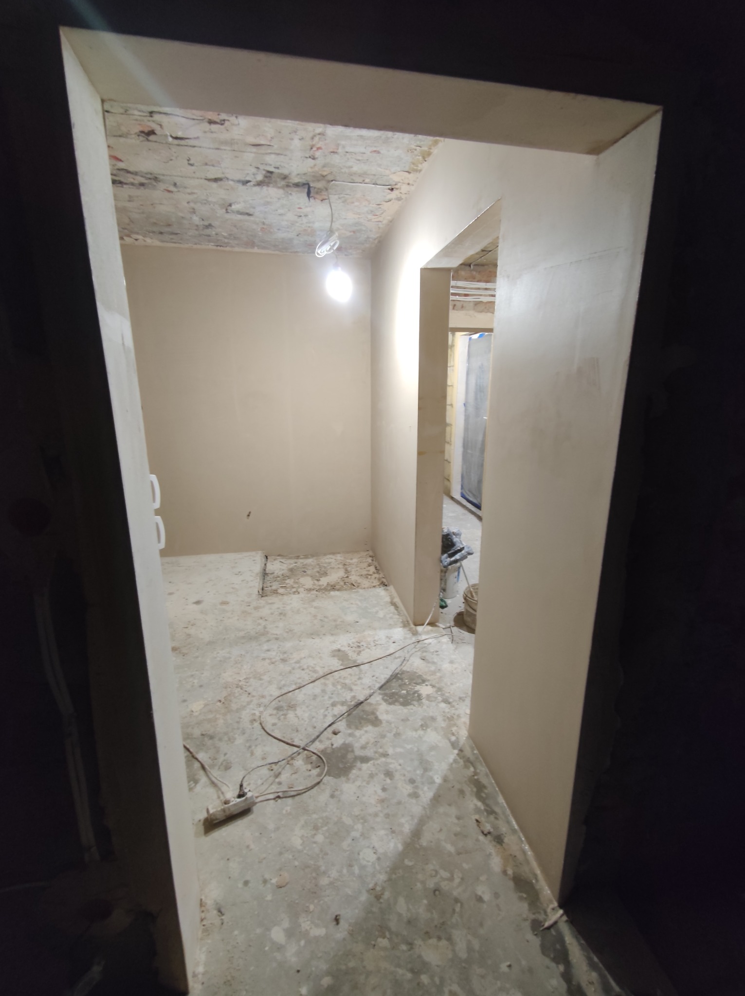 Wnętrze w trakcie remontu: świeżo położone gładzie na ścianach, surowy beton na podłodze, widoczne kable elektryczne i niedokończony sufit z wiszącą żarówką.