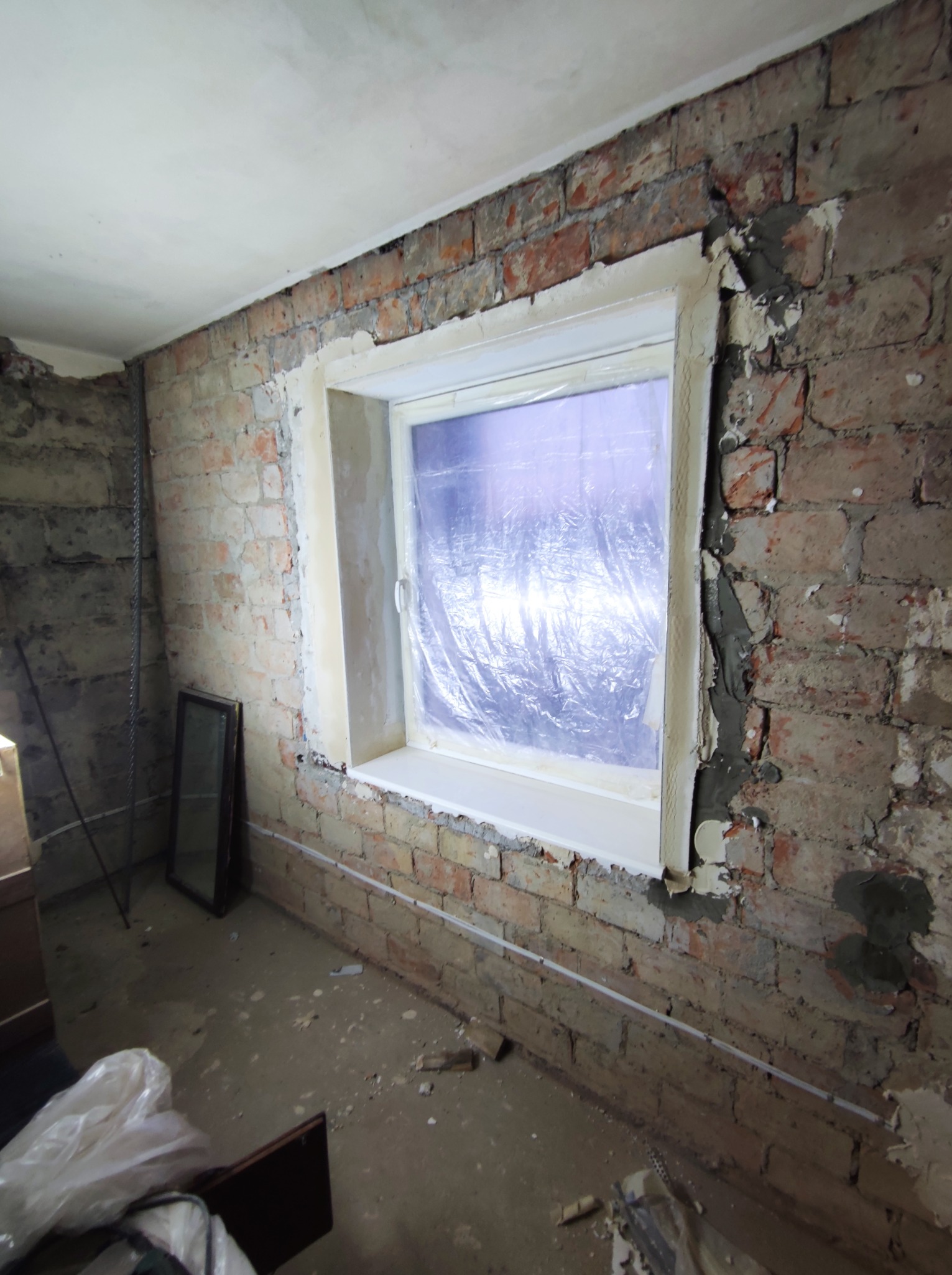 Pokój w trakcie remontu: odsłonięta cegła na ścianach, okno zabezpieczone folią, przygotowane do dalszych prac wykończeniowych.