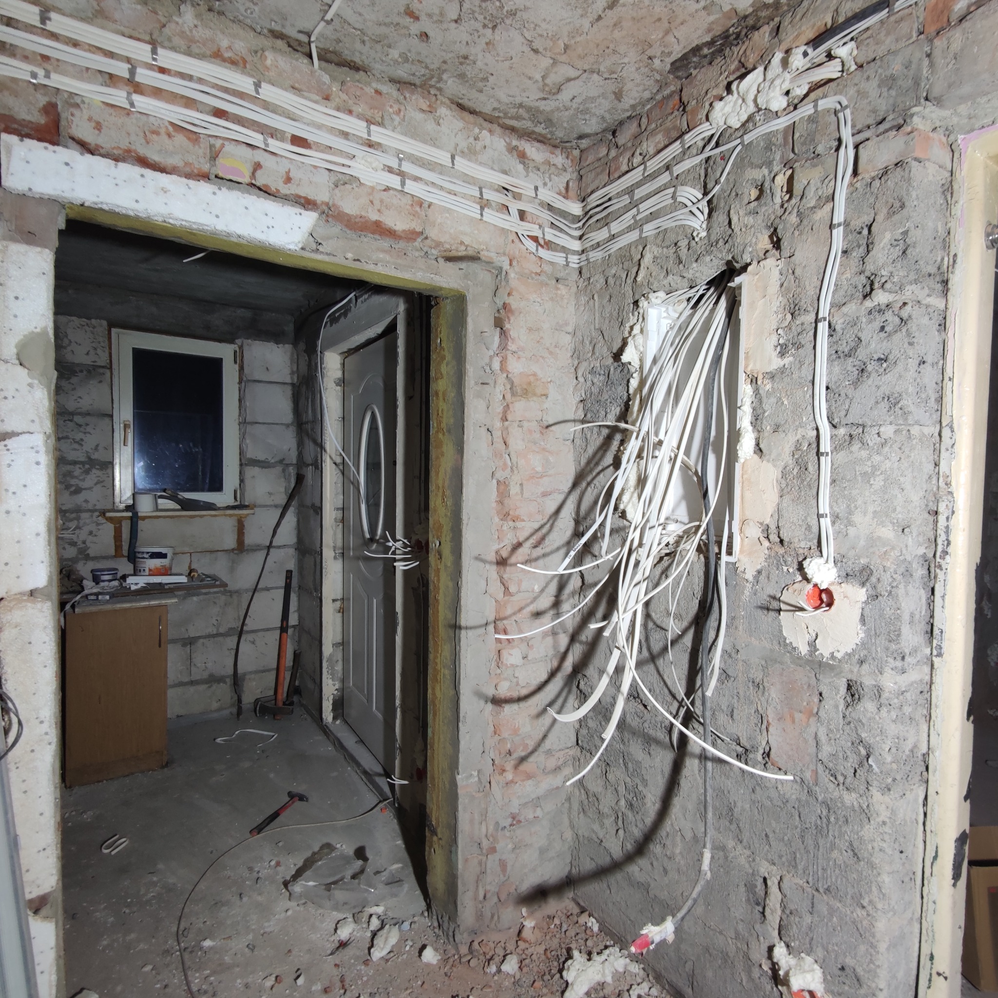 Wnętrze w trakcie remontu: odsłonięta cegła, instalacja elektryczna w peszlach, ślady po kuciu, pianka montażowa, widoczne przewody w ścianie, okno i drzwi w tle.