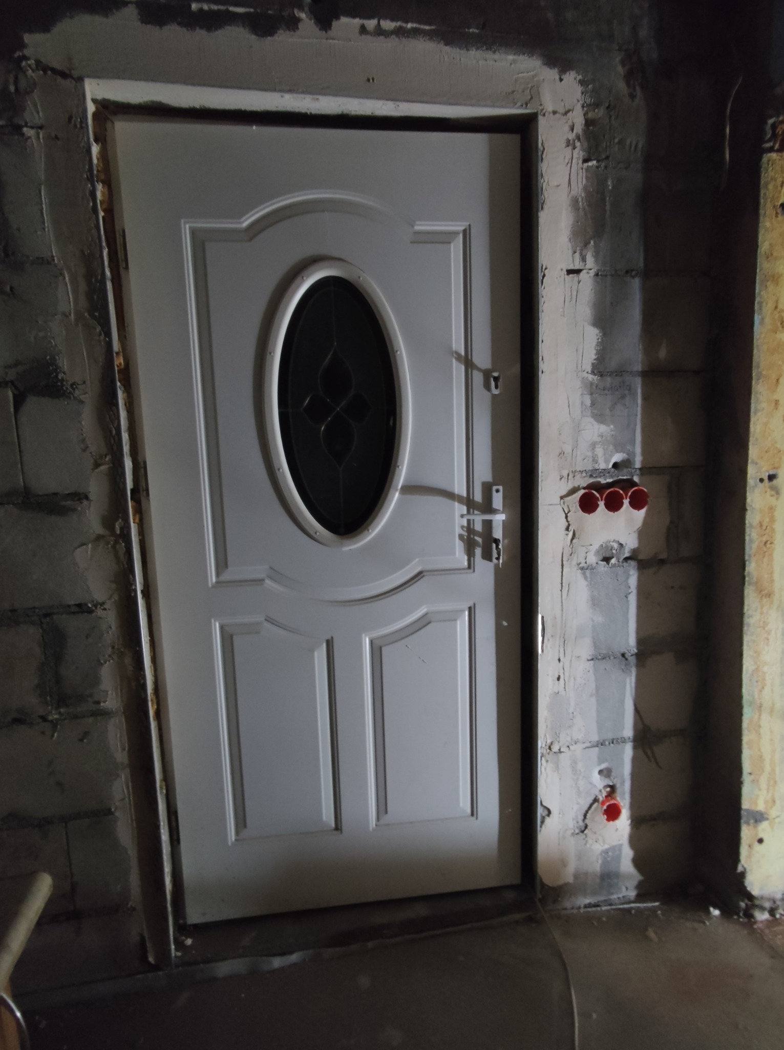 Białe drzwi wejściowe z owalnym przeszkleniem, osadzone w surowym otworze ściennym z widocznymi blokami betonowymi i instalacją elektryczną z czerwonymi rurkami, w trakcie prac wykończeniowych.