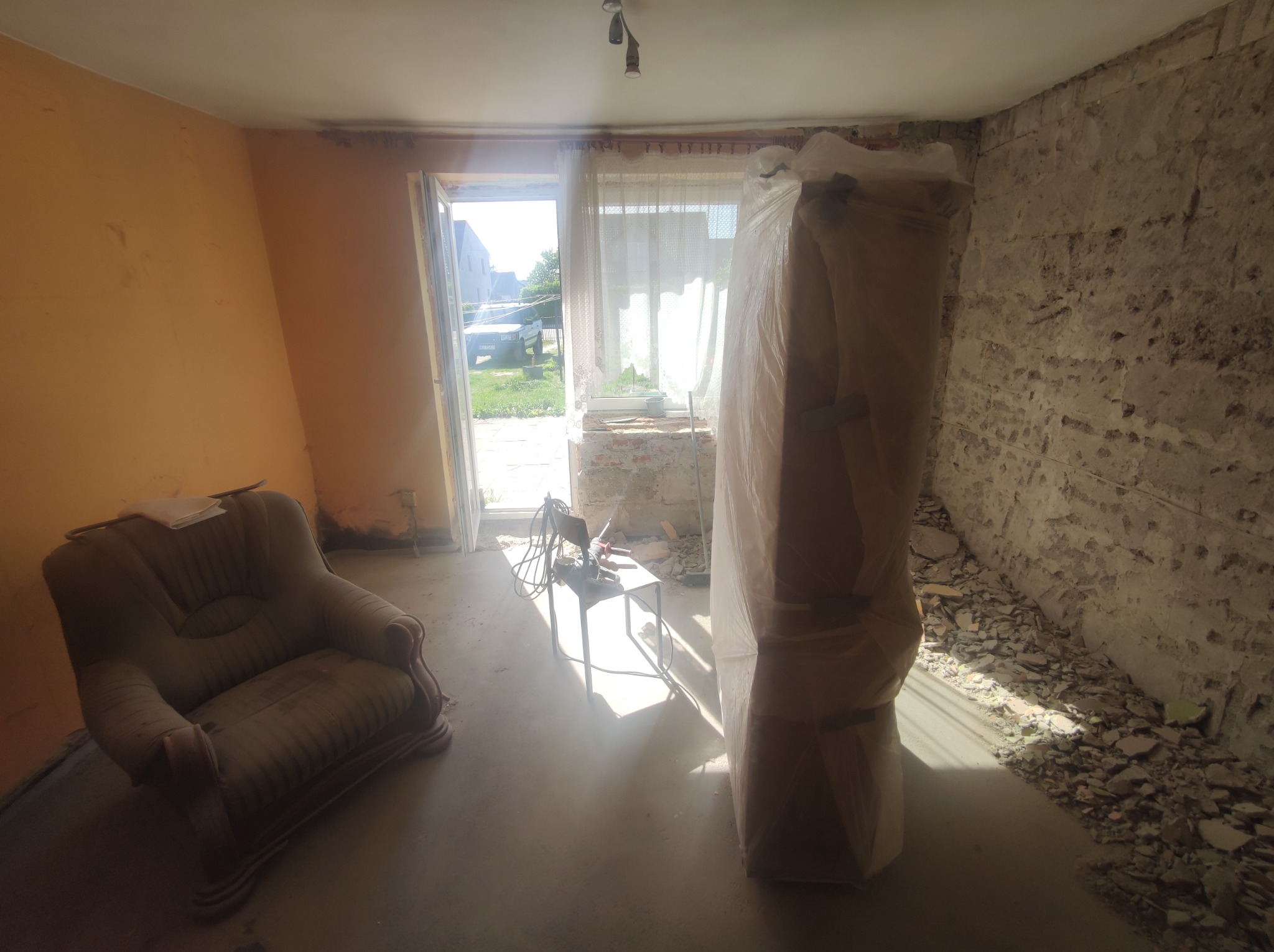 Pokój w trakcie remontu: pomarańczowa ściana, odsłonięta cegła, okno z widokiem na zewnątrz, fotel i mebel zabezpieczony folią.