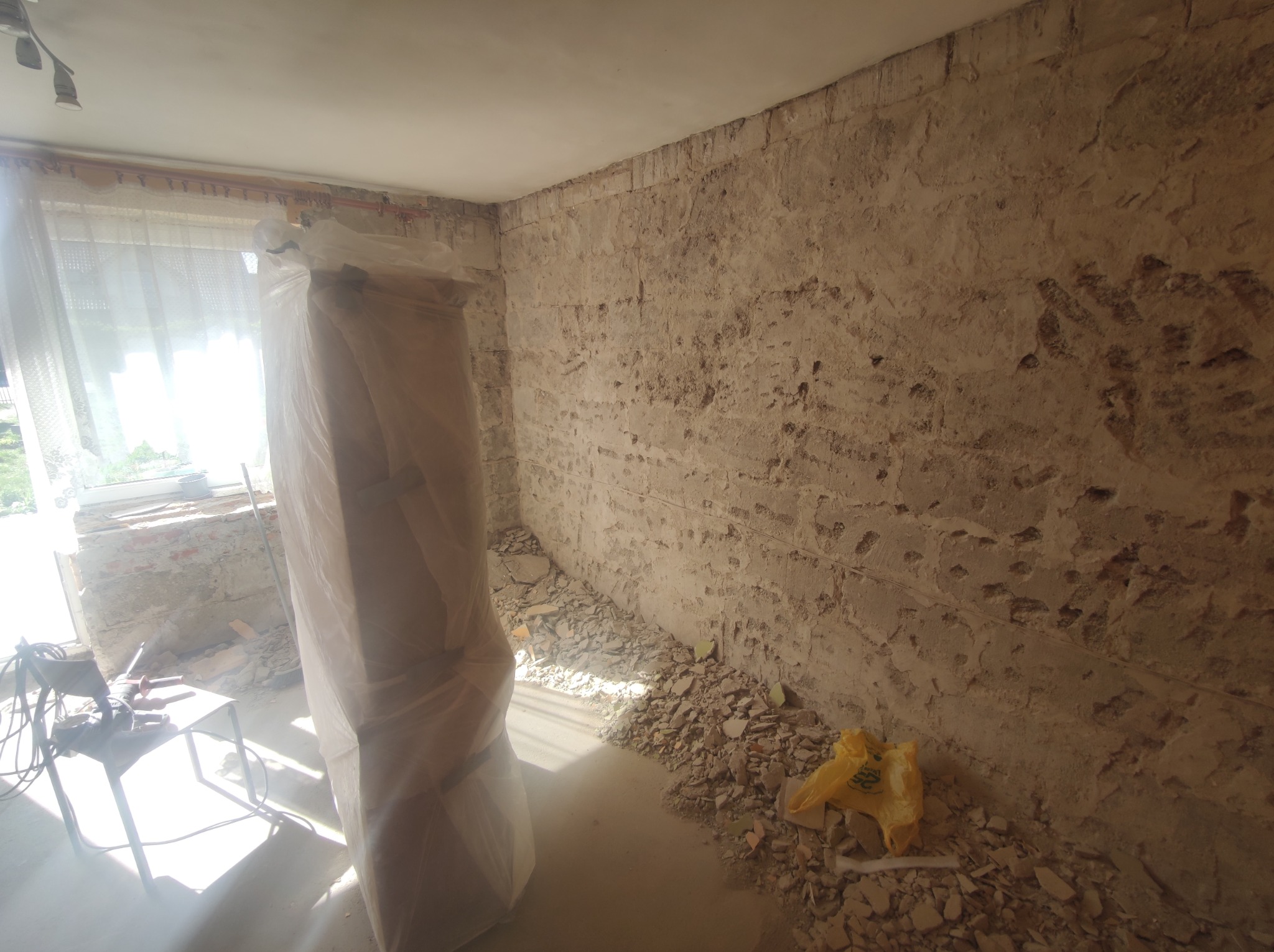 Pokój w trakcie skuwania tynku ze ścian, widoczne narzędzia na stole, zabezpieczone folią drzwi i gruz na podłodze.