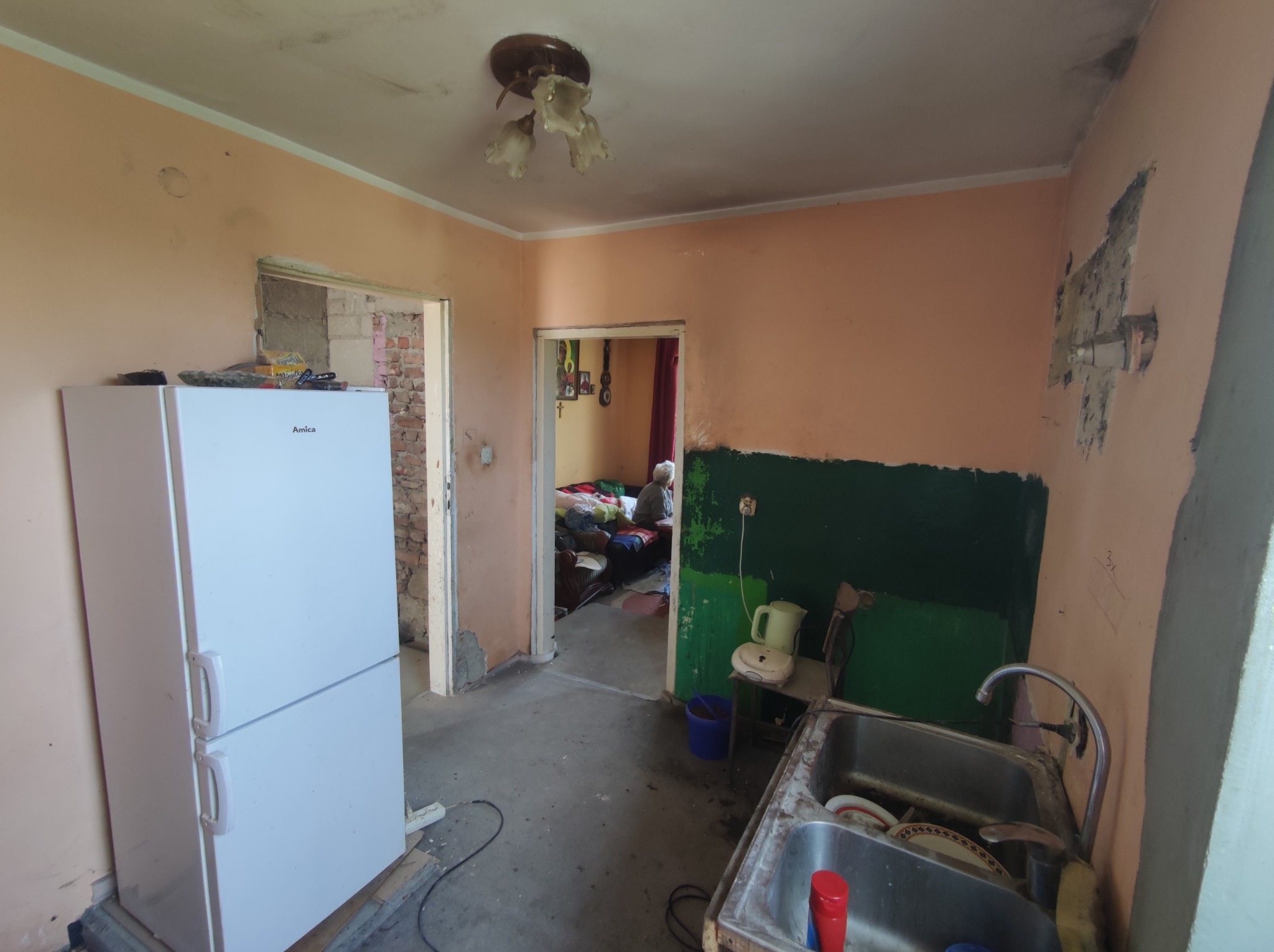 Kuchnia w trakcie remontu: ściany częściowo pomalowane na zielono, pozostałe w odcieniach brzoskwini, widoczne ślady po wcześniejszych pracach. Stara instalacja wodna z zardzewiałym zlewem. W tle...