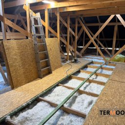 Termool - Ocieplenie poddasza wełną mineralną, widoczne krokwie, płyty OSB i drabina. Prace w toku, oświetlenie halogenowe. Widoczny fragment logo firmy.