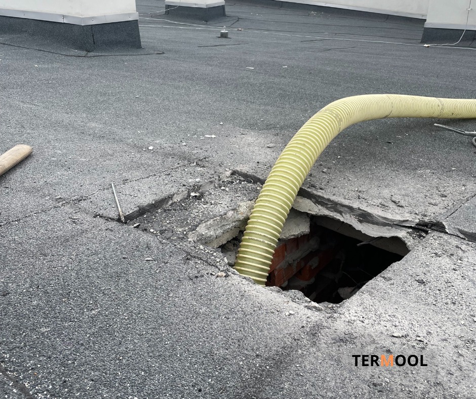 Żółty wąż wpuszczony w otwór w płaskim dachu podczas prac izolacyjnych, widoczne cegły w otworze, na dachu widoczne pozostałości gruzu i narzędzia.