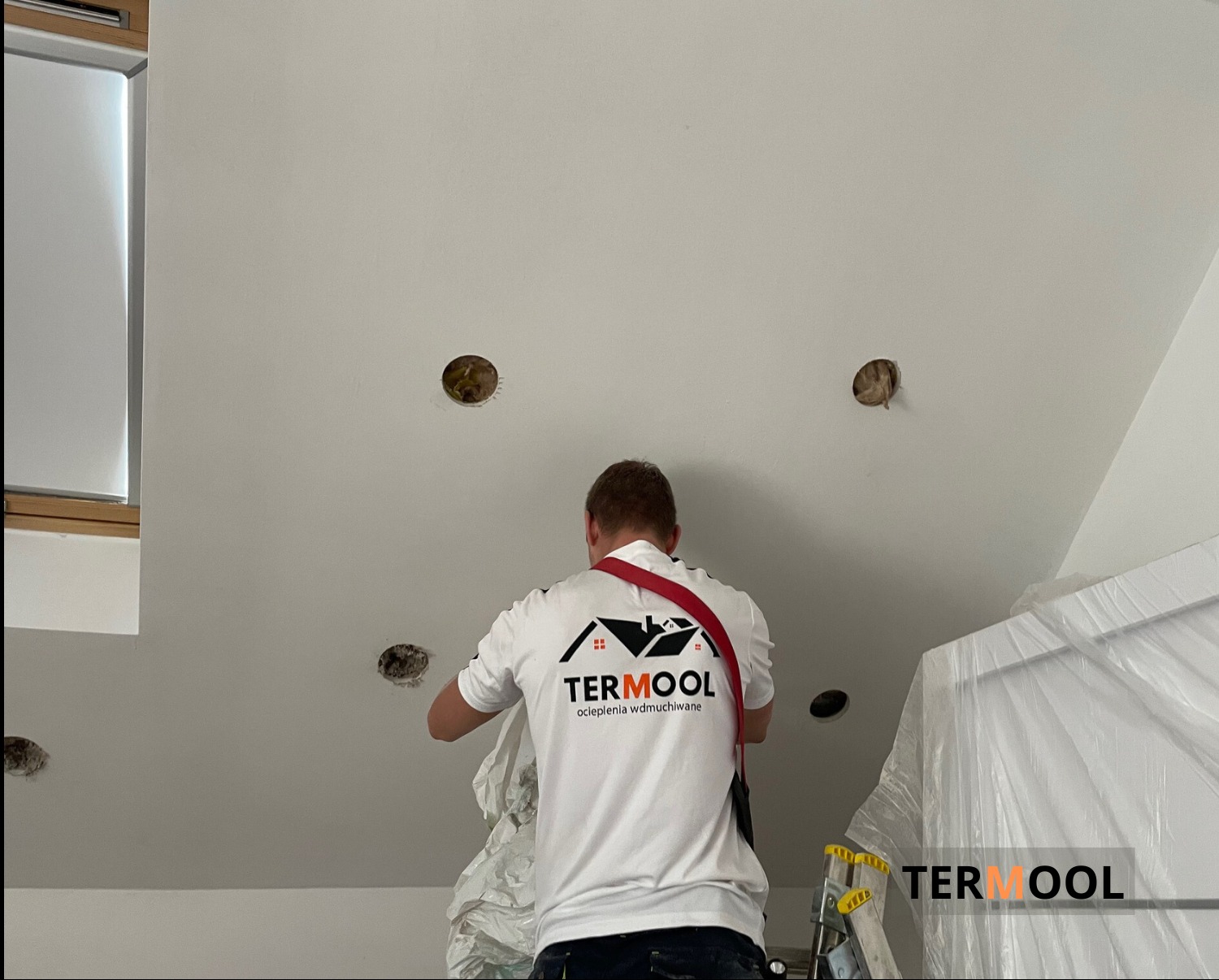 Pracownik firmy Termool w trakcie prac przygotowawczych do ocieplania poddasza metodą wdmuchiwania, widoczne otwory w suficie oraz logo firmy na koszulce.