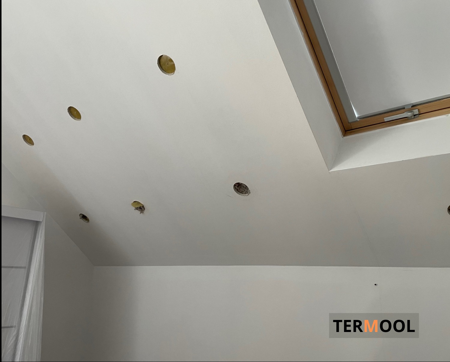 Białe poddasze z widocznymi otworami na oświetlenie punktowe przed ociepleniem, okno dachowe z drewnianą ramą w rogu, częściowo zakryte drzwi po lewej stronie, logo firmy Termool w prawym dolnym rogu.
