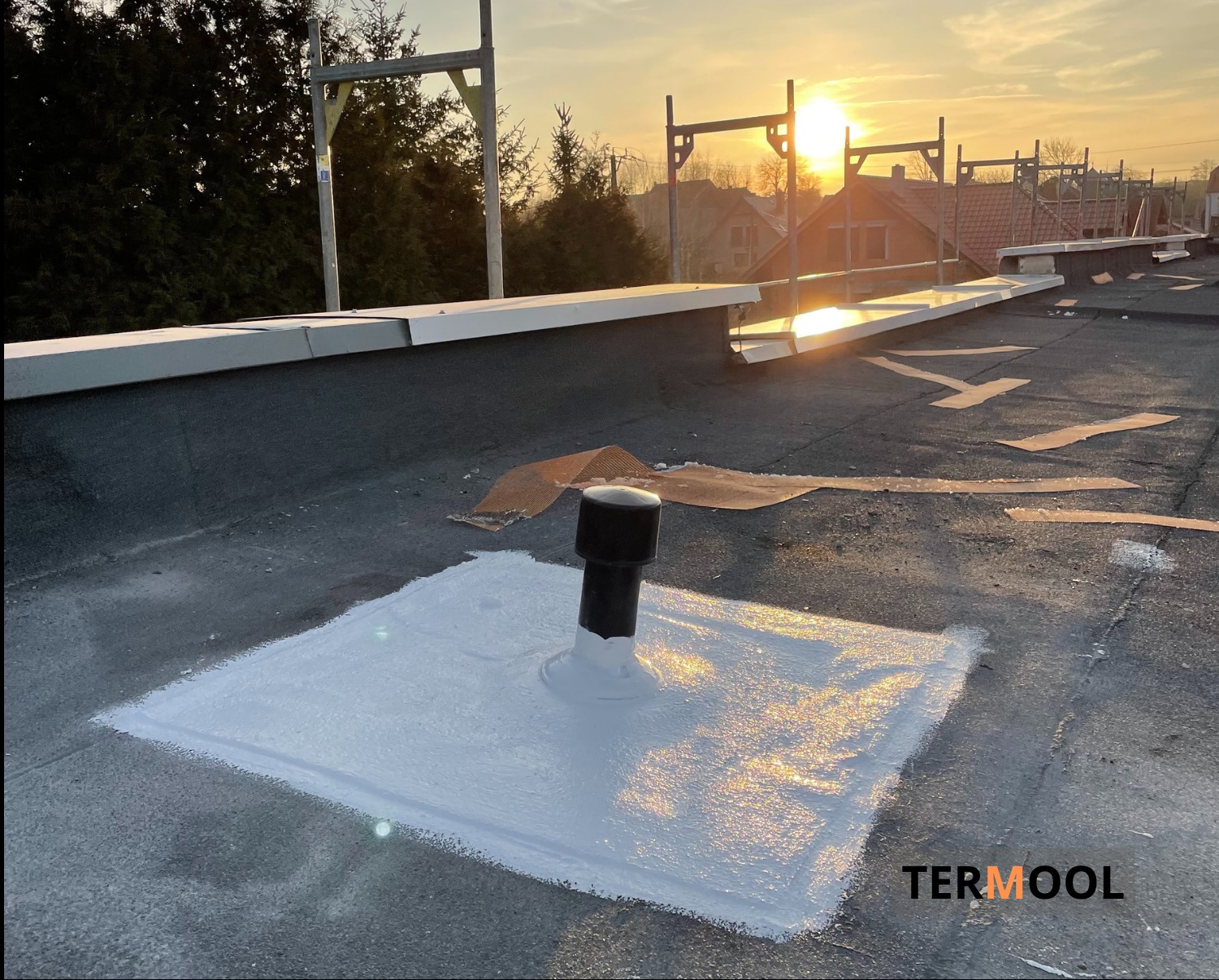 Płaski dach pokryty bitumiczną membraną, z uszczelnionym kominkiem wentylacyjnym świeżą warstwą białej powłoki hydroizolacyjnej, w tle zachodzące słońce i rusztowania.