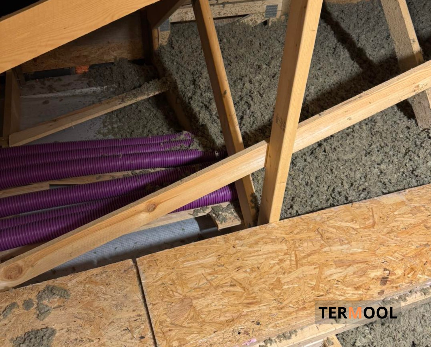 Poddasze z drewnianą konstrukcją, wypełnione szarą wełną izolacyjną i fioletowymi rurami wentylacyjnymi. Widoczne płyty OSB i logo firmy w rogu.