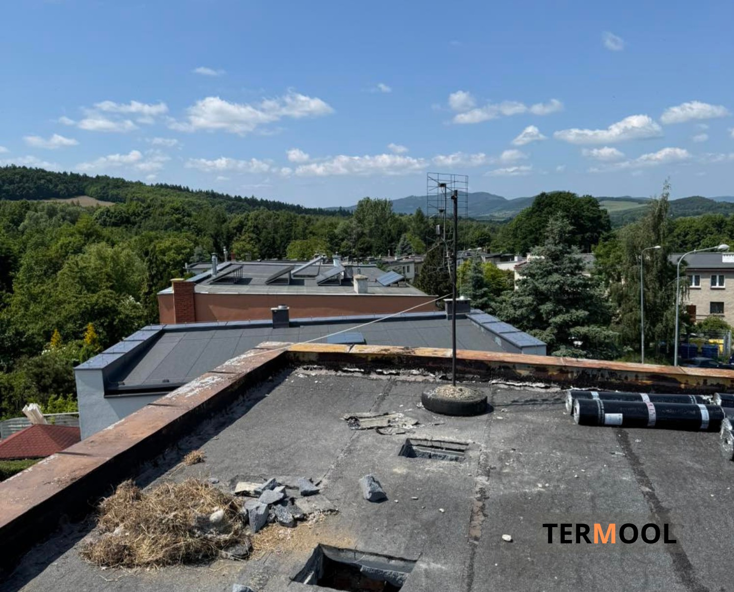 Niedokończony płaski dach z widocznymi uszkodzeniami, gruzem i rolkami papy. W tle zabudowa miejska i zalesione wzgórza pod błękitnym niebem.