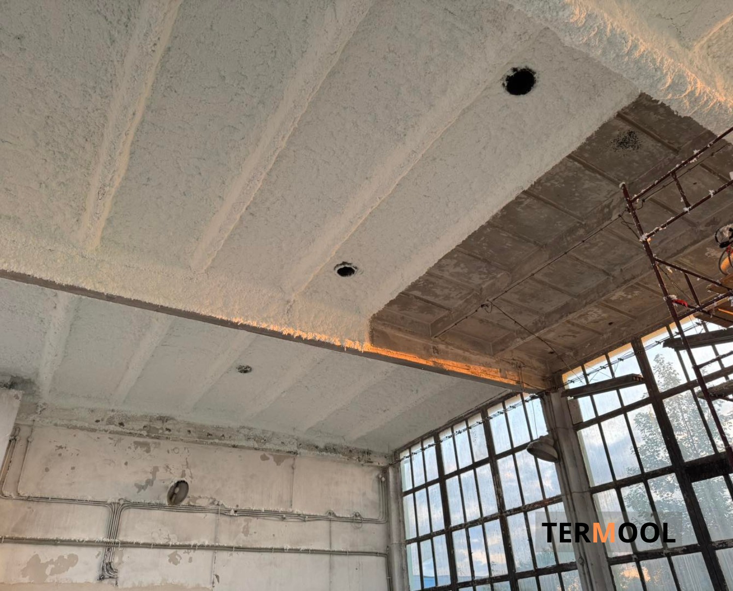 Poddasze z częściowo nałożoną pianką PUR, widoczne elementy konstrukcyjne i okna. Fragment rusztowania w prawym górnym rogu. Oświetlenie naturalne.