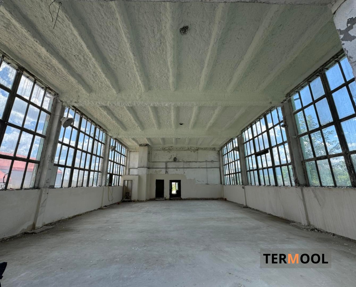 Poddasze przemysłowe po ociepleniu pianką PUR, widoczne okna z licznymi szybami i równomierna warstwa izolacji na suficie. Duża przestrzeń, surowy beton na podłodze.