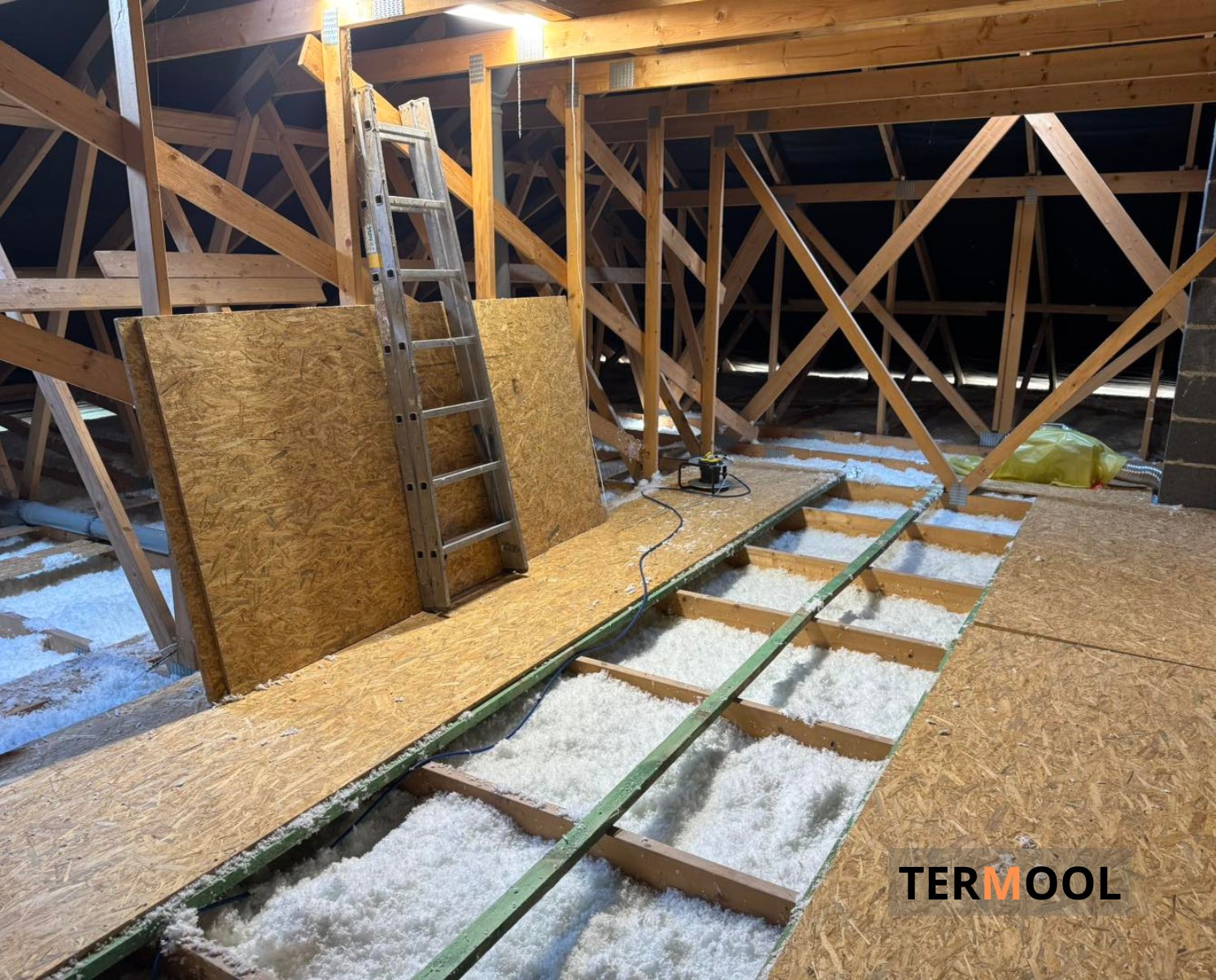 Ocieplenie poddasza wełną mineralną, widoczne krokwie, płyty OSB i drabina. Prace w toku, oświetlenie halogenowe. Widoczny fragment logo firmy.