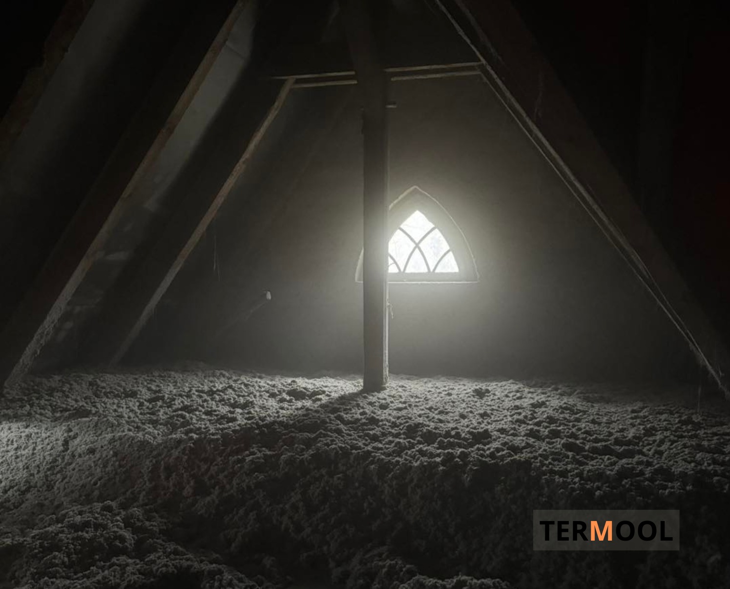 Poddasze z oknem w stylu gotyckim, wypełnione szarą wełną izolacyjną. Widoczne drewniane elementy konstrukcyjne i logo firmy w prawym dolnym rogu.