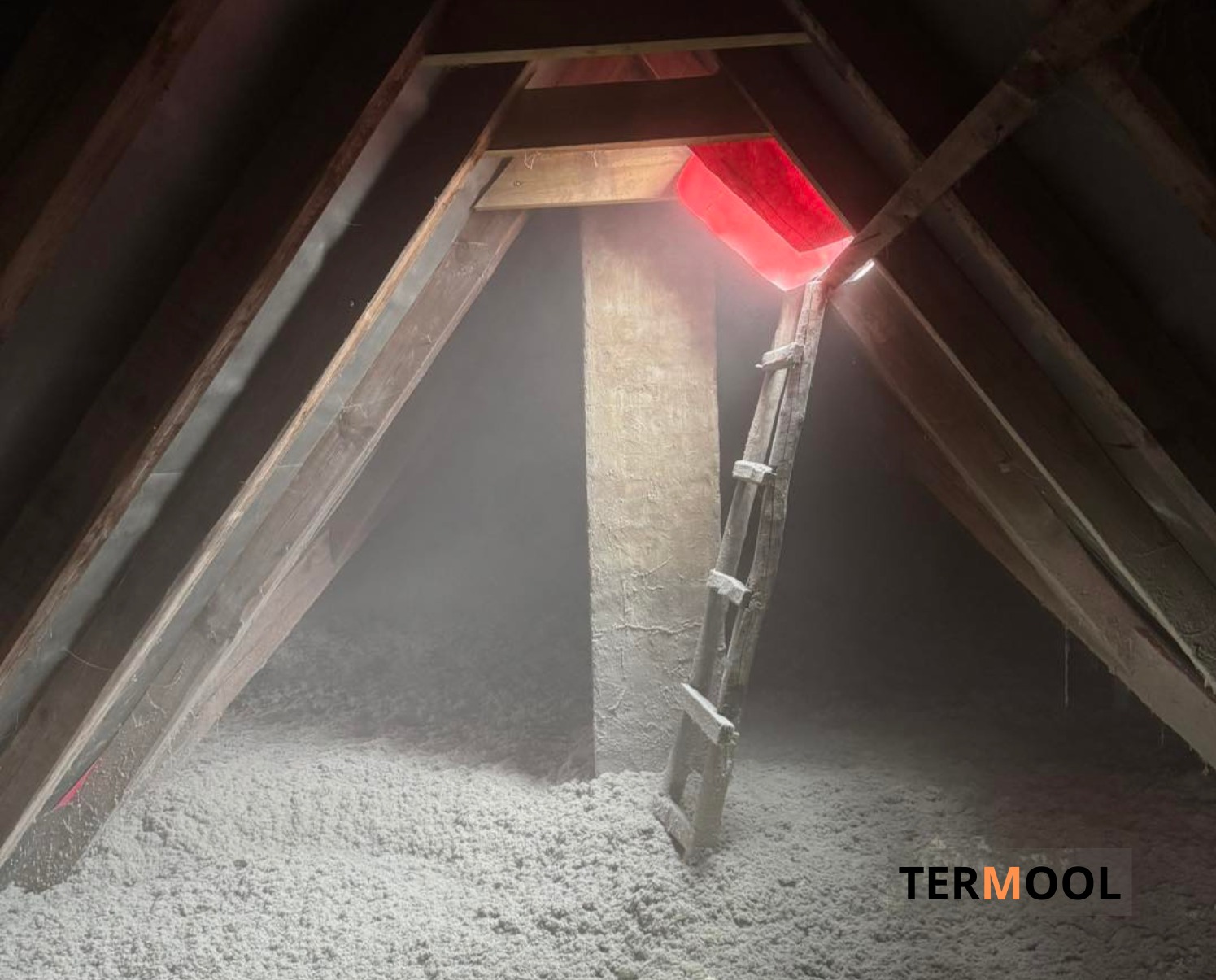 Poddasze po ociepleniu wełną celulozową, widoczne drewniane krokwie, drabina i czerwone światło wpadające z zewnątrz. Widoczny znak firmowy w prawym dolnym rogu.