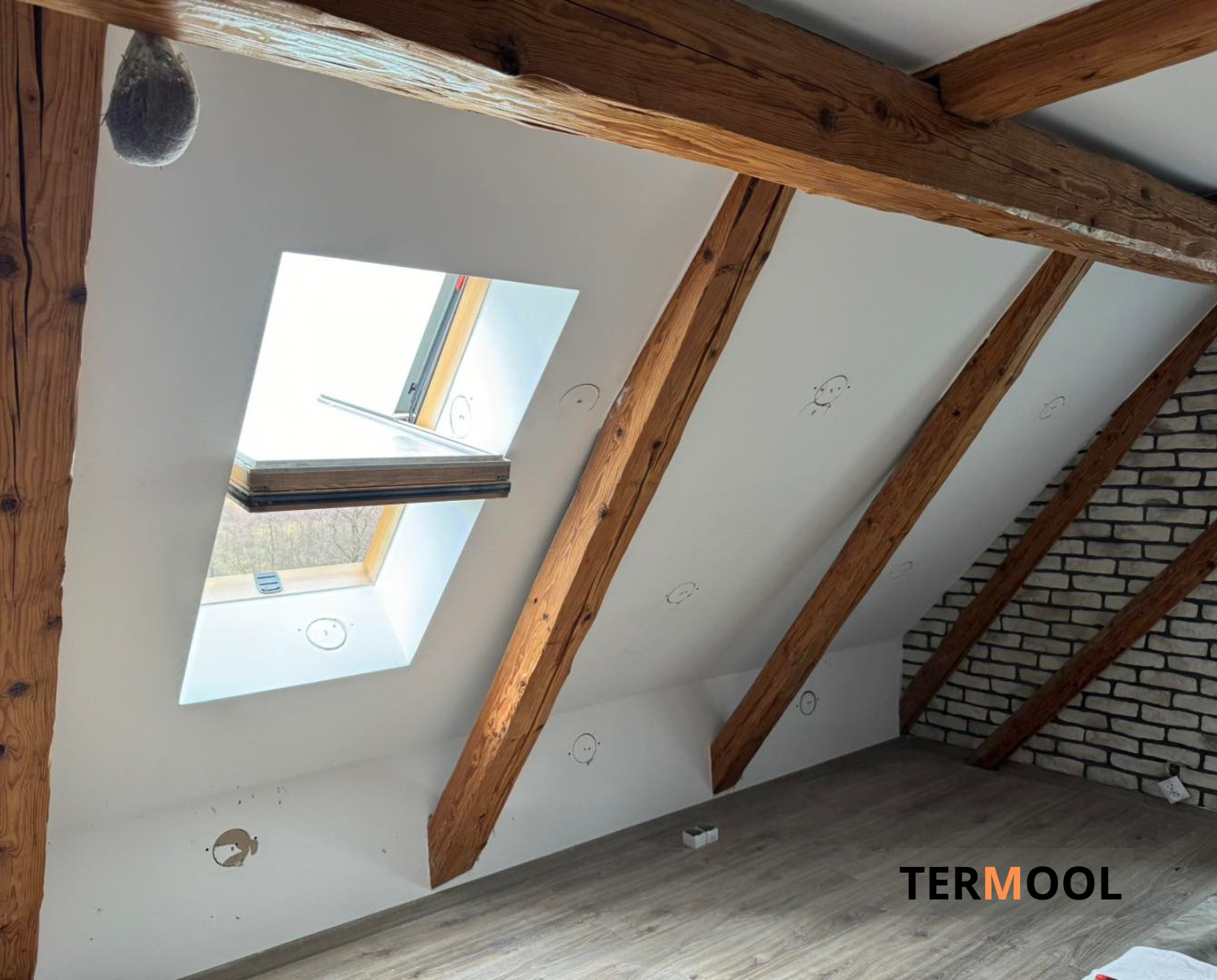 Poddasze po ociepleniu z widocznymi drewnianymi belkami, oknem dachowym i fragmentem ściany z cegły. Widoczne ślady po instalacji elektrycznej.