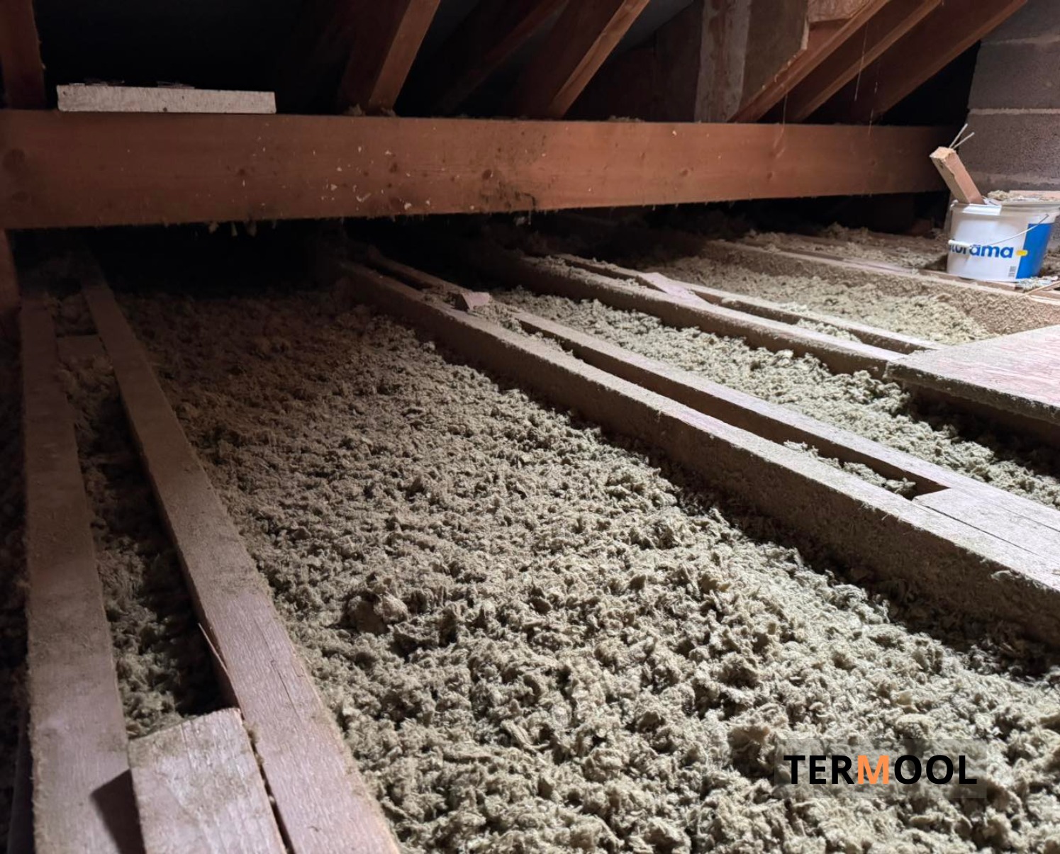 Poddasze z widoczną warstwą izolacji z wełny mineralnej między drewnianymi belkami. W tle wiadro z farbą. Ujęcie z dołu, ukazujące strukturę materiału.