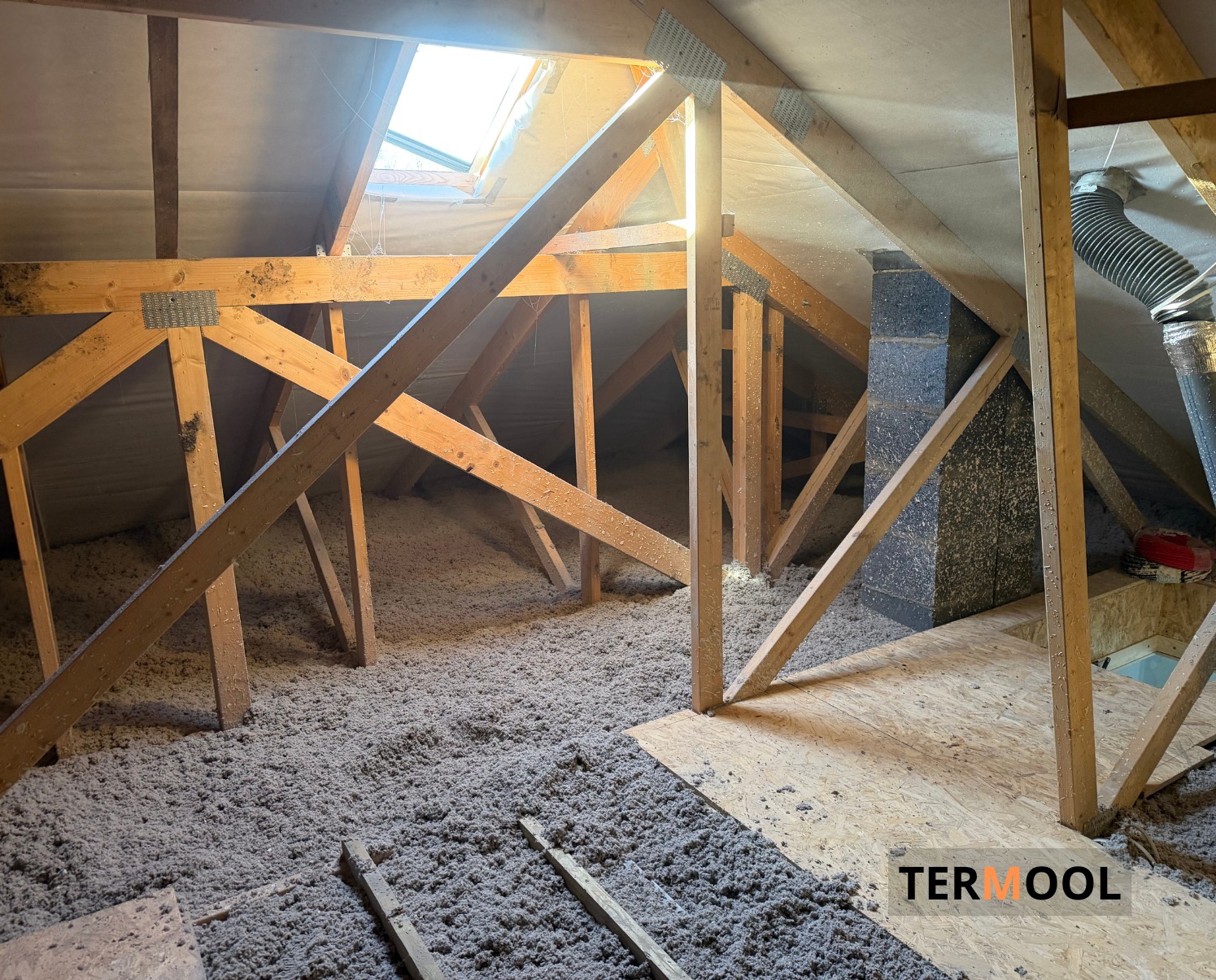 Poddasze z drewnianą konstrukcją i oknem dachowym, wypełnione szarą izolacją z włókien. Widoczny komin i fragment podłogi z płyt OSB. Ujęcie z dołu.