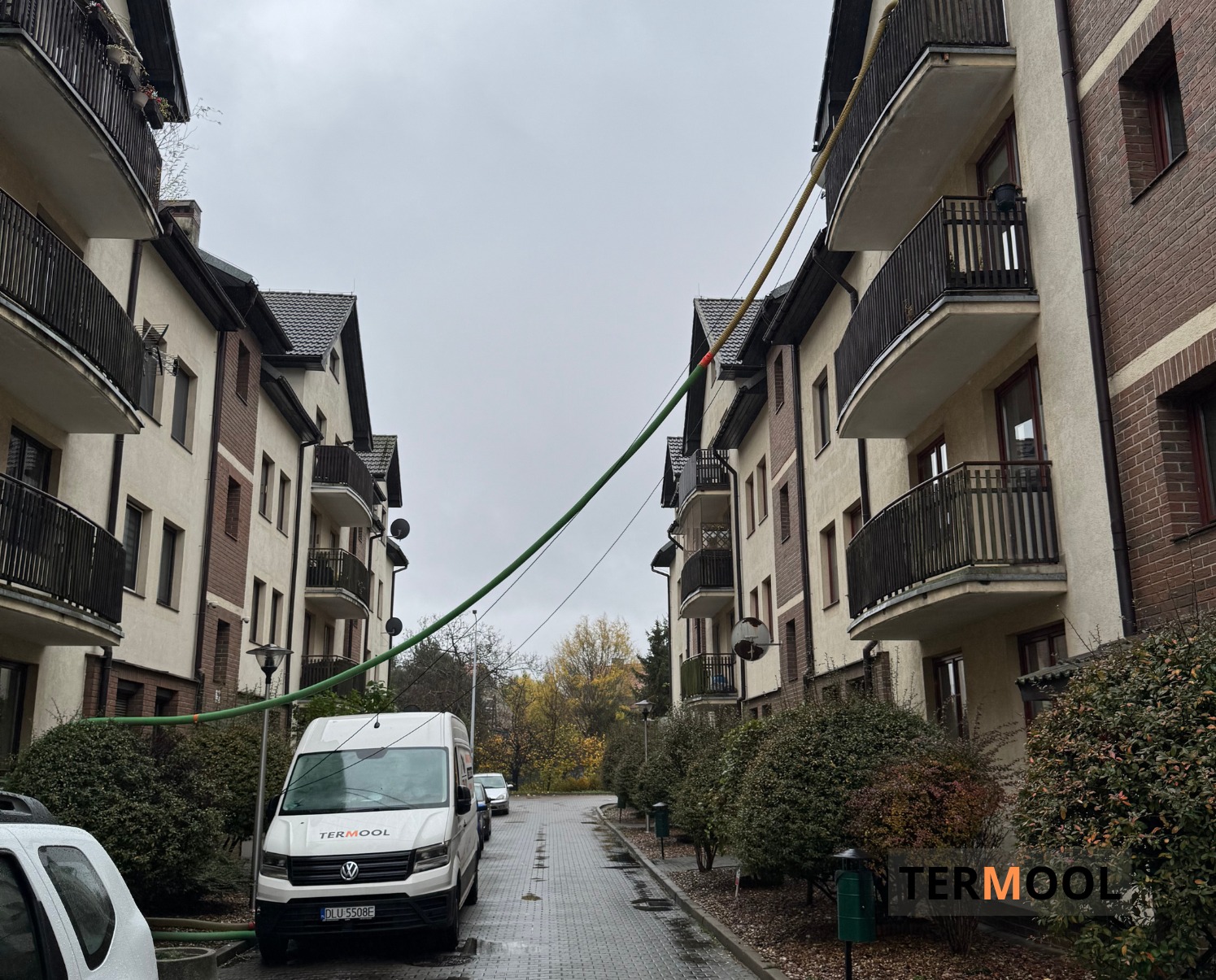 Ocieplanie pianką PUR bloku mieszkalnego. Wąż z pianką prowadzony z samochodu dostawczego Termool. Widok na budynek z balkonami i elewacją w odcieniach beżu i brązu.