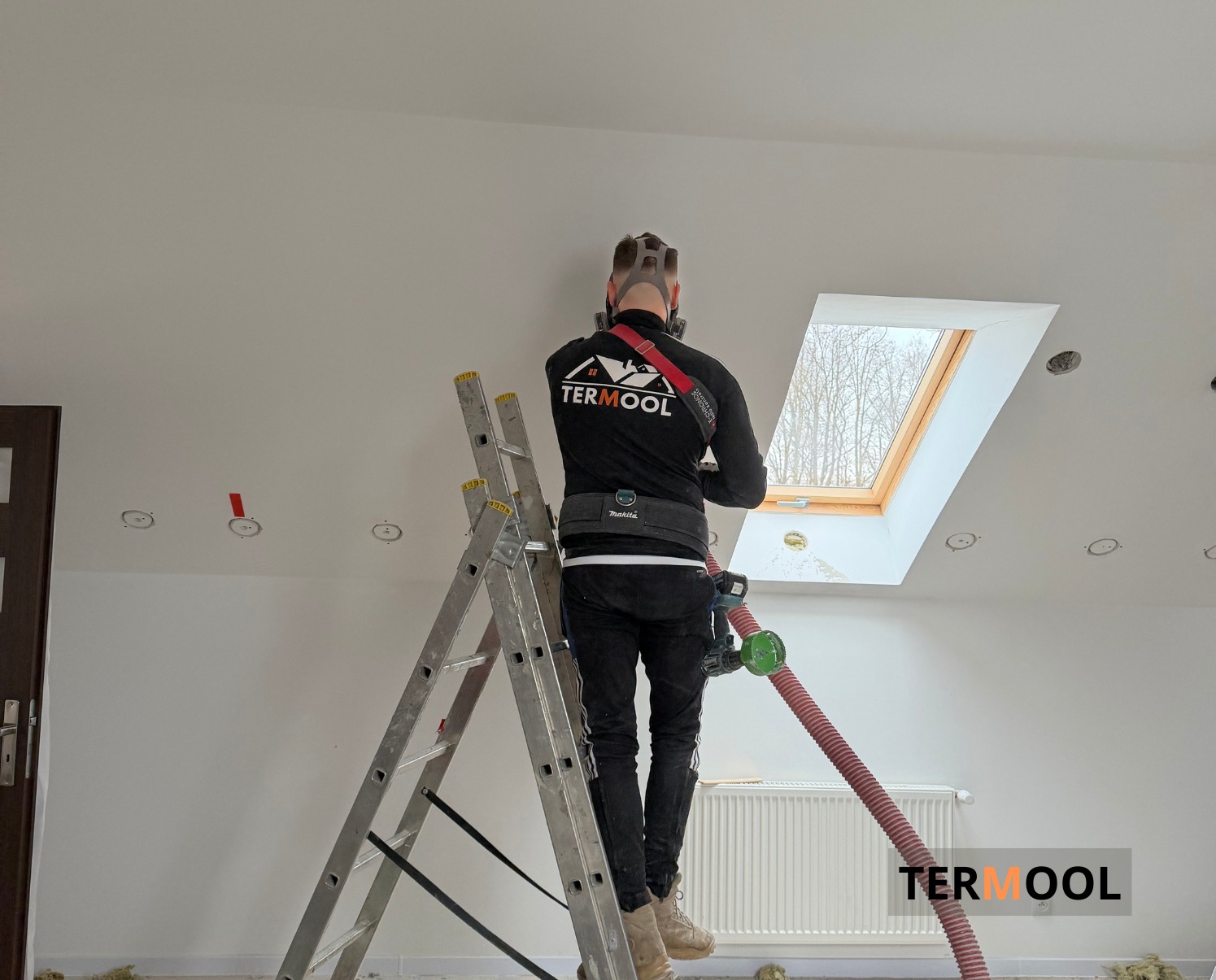 Pracownik w masce na drabinie, aplikuje pianę PUR na poddaszu z oknem dachowym. Widoczny sprzęt do natrysku i logo firmy na ubraniu. Białe ściany i sufit.