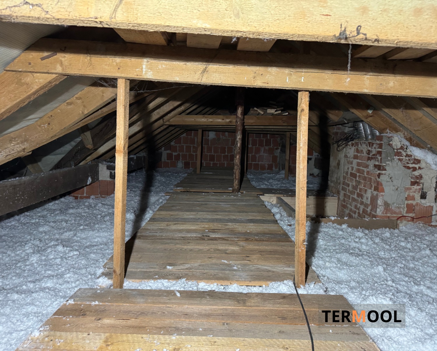 Poddasze ocieplone białą wełną mineralną, widoczne drewniane elementy konstrukcyjne i ceglany komin. Fragment drewnianej ścieżki ułatwia poruszanie się po ociepleniu.