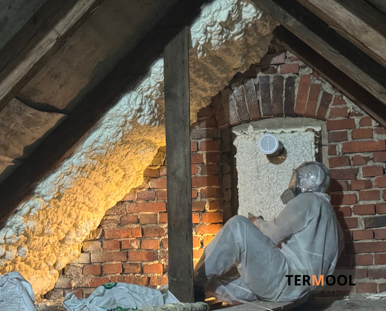 Poddasze ocieplone pianką PUR, widoczna ceglana ściana i pracownik w kombinezonie ochronnym. Oświetlenie podkreśla strukturę pianki i cegieł.