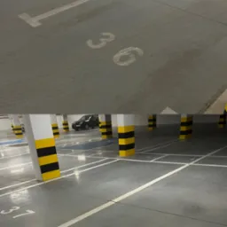 Wnętrze parkingu podziemnego z betonową podłogą, białymi liniami wyznaczającymi miejsca parkingowe z numerami 36 i 37, oraz kolumnami w kolorze białym z czarno-żółtymi pasami. Widoczne ślady opon...