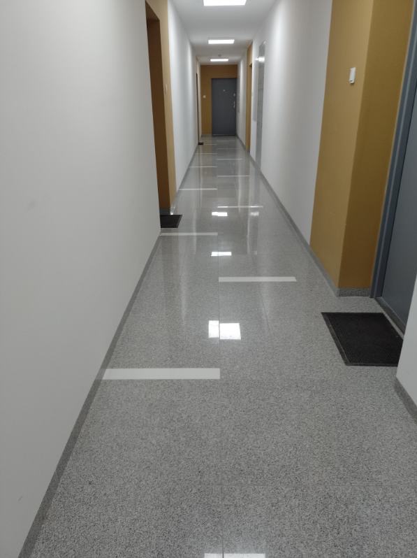 Długi korytarz w budynku biurowym z błyszczącą, szarą podłogą z terazzo i białymi pasami, oświetlony lampami sufitowymi, z widocznymi drzwiami i kolumnami w kolorze beżowym.