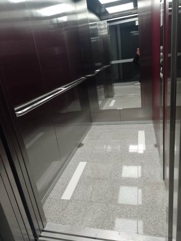 Wnętrze windy z metalowymi ścianami, poręczą i jasnoszarą podłogą w plamy, z białymi paskami antypoślizgowymi.