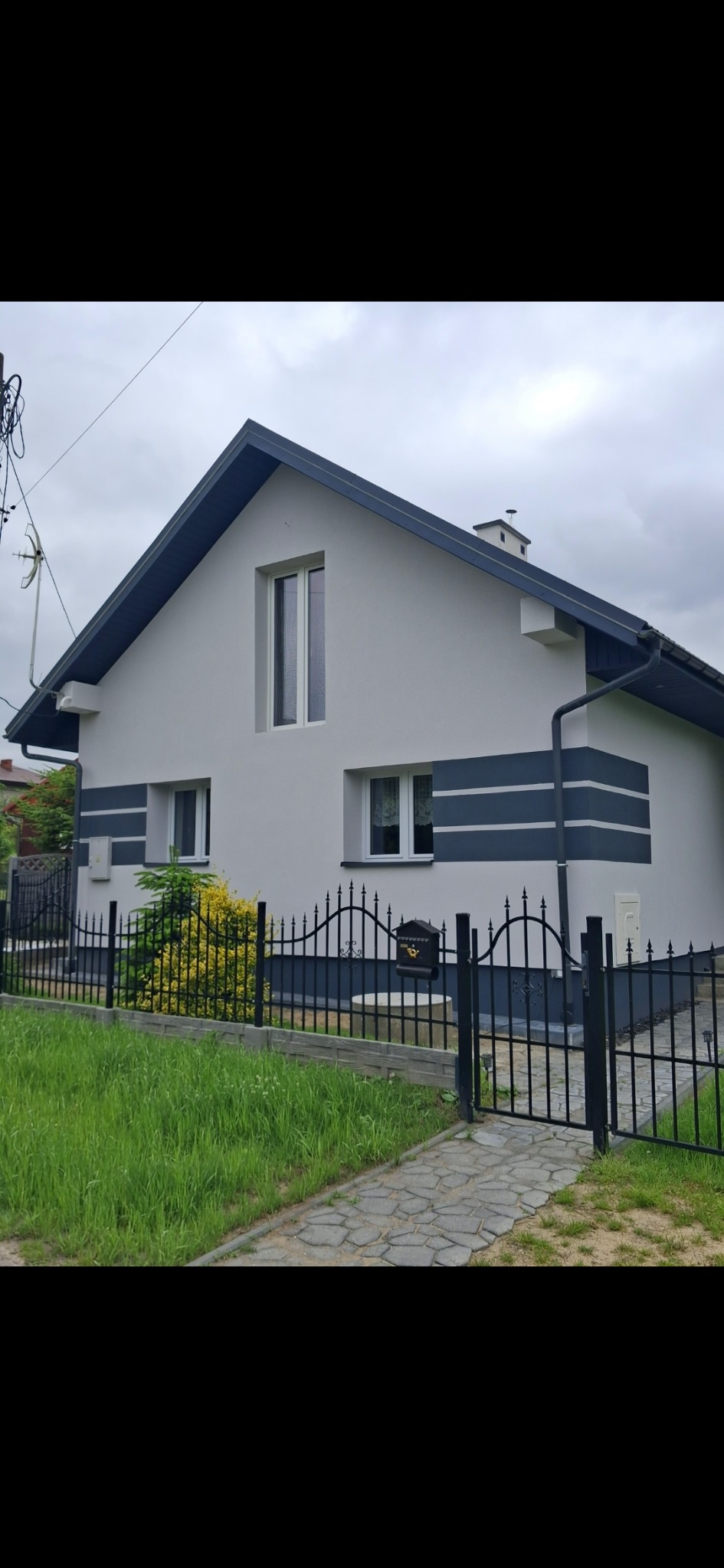 Dwukondygnacyjny dom z szarą elewacją, ciemnymi pasami i dachem. Widoczne okna, rynna, metalowe ogrodzenie i skrzynka pocztowa. Pochmurne niebo w tle.