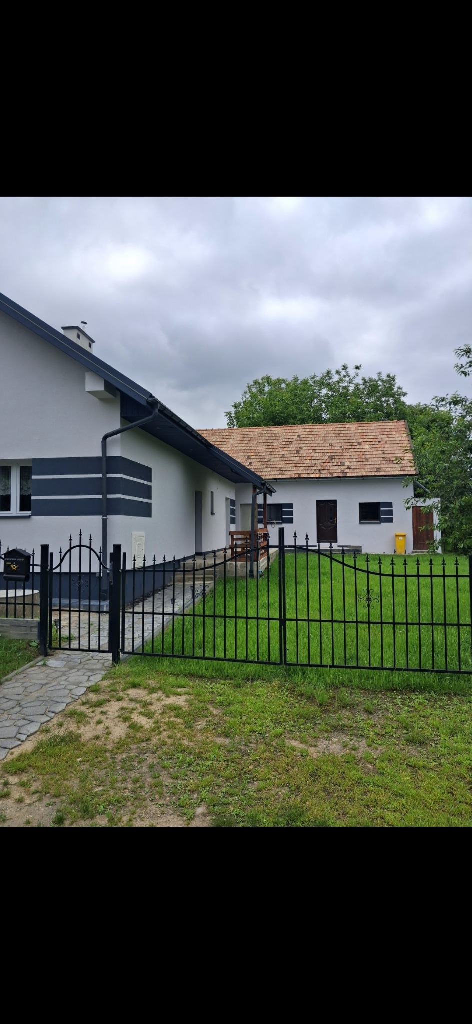 Dwa budynki połączone, z nową, jasną elewacją i ciemnymi akcentami, widoczne zza czarnego, kutego ogrodzenia na tle zielonego trawnika i pochmurnego nieba.