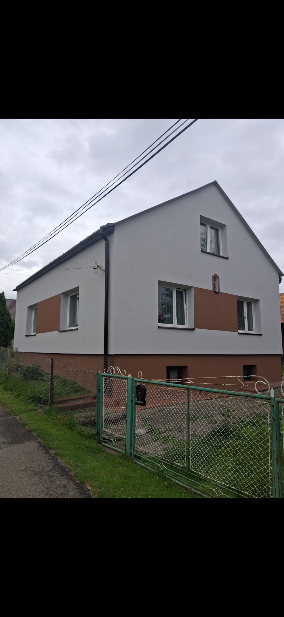 Dwukondygnacyjny dom z nową, dwukolorową elewacją (szary i brązowy) i białymi oknami, widziany zza zielonego ogrodzenia siatkowego w pochmurny dzień.