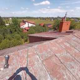 Łukasz JACHIMOWICZ - Czerwony dach z łuszczącą się farbą, widoczny komin z cegły, narzędzie akumulatorowe i liny zabezpieczające na tle wiejskiego krajobrazu.