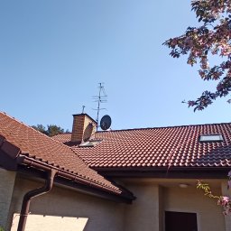 Łukasz JACHIMOWICZ - Czerwona dachówka ceramiczna na dachu domu z kominem, anteną satelitarną i kwitnącym drzewem w prawym górnym rogu.