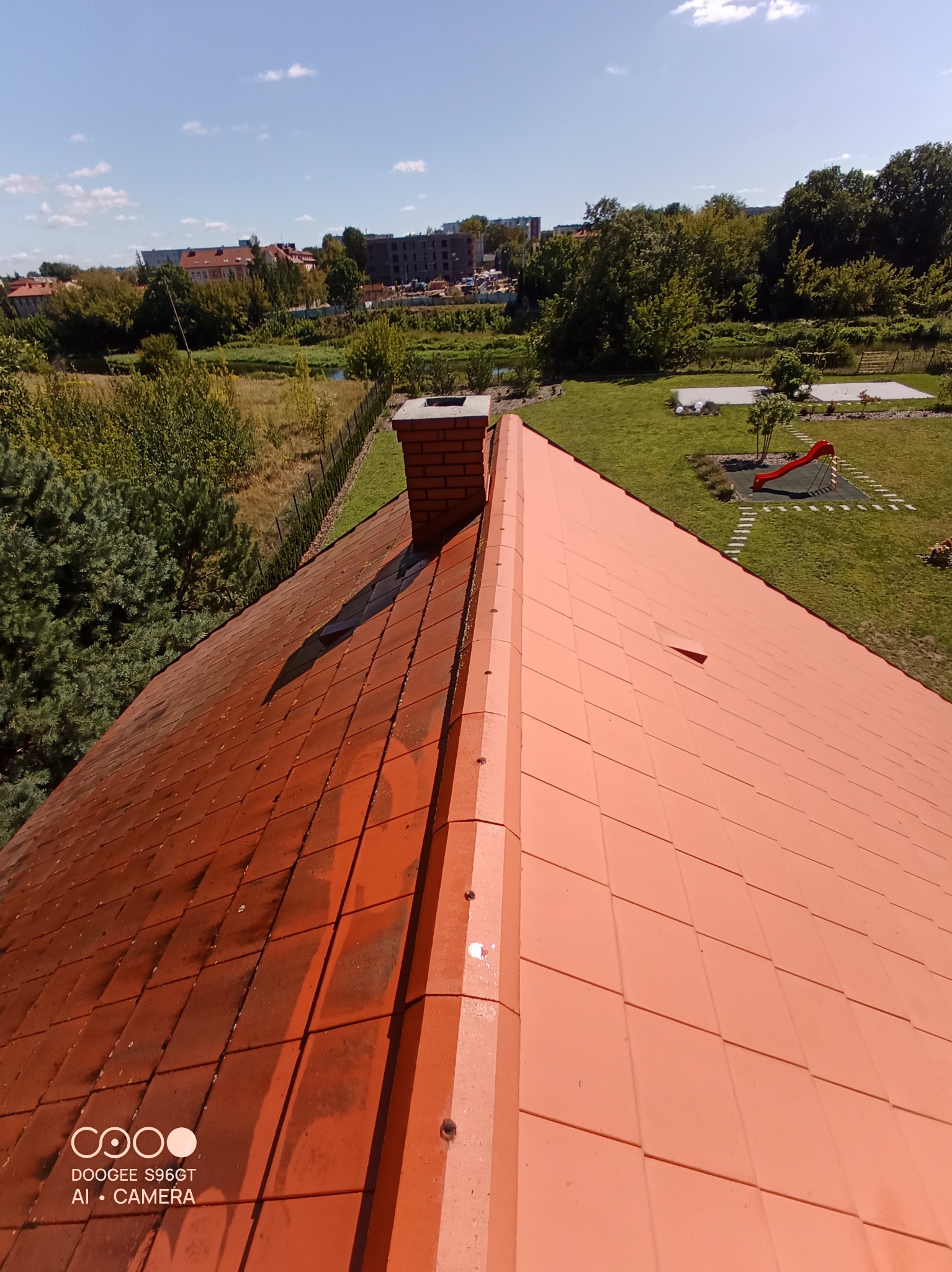 Częściowo wyczyszczony dach z czerwonej dachówki ceramicznej, widoczny komin z cegły i otoczenie z zielenią, ukazujące różnicę w wyglądzie przed i po czyszczeniu.