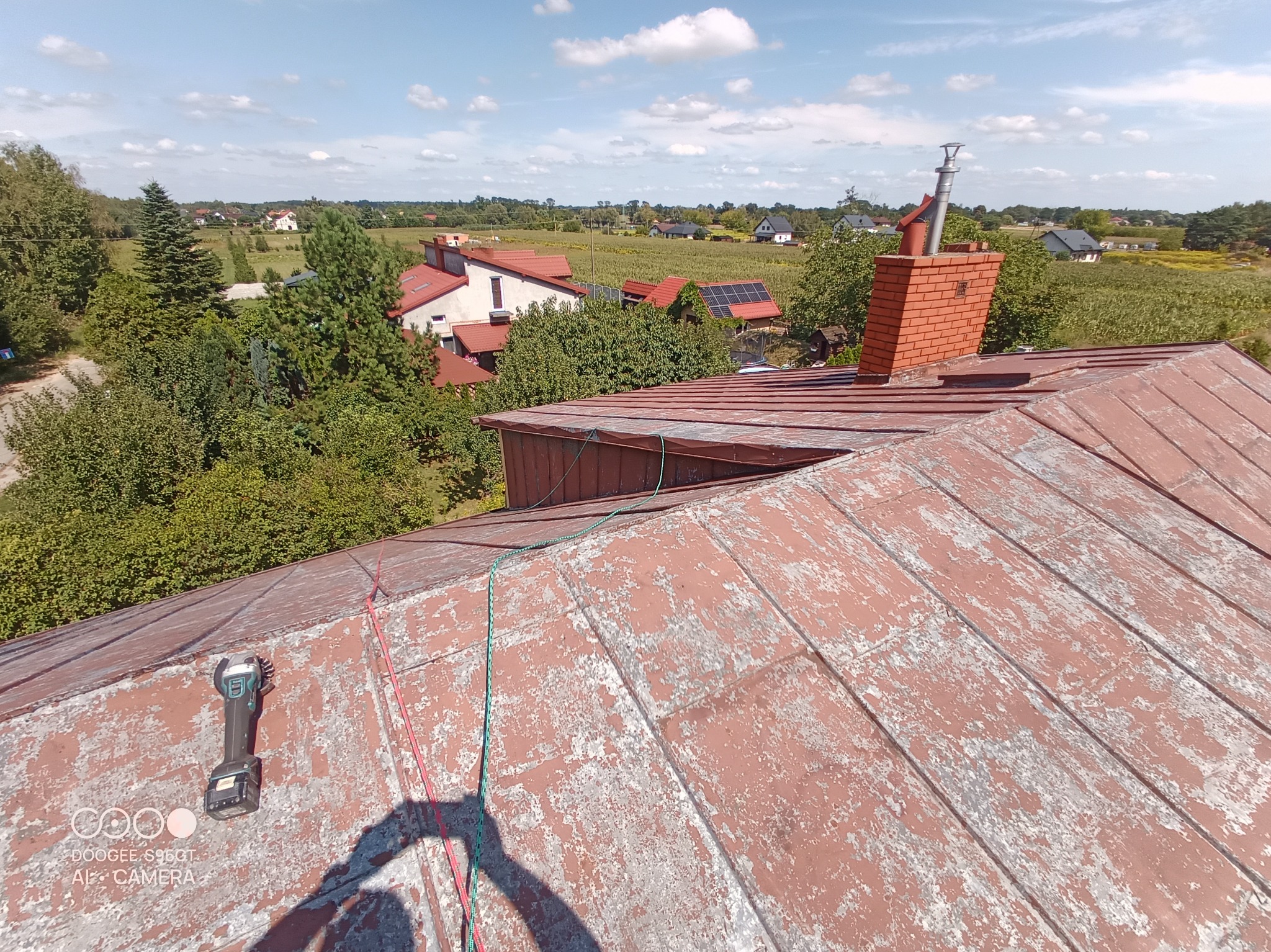 Czerwony dach z łuszczącą się farbą, widoczny komin z cegły, narzędzie akumulatorowe i liny zabezpieczające na tle wiejskiego krajobrazu.