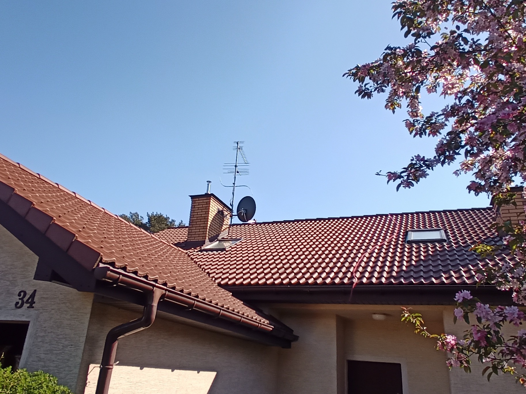 Czerwona dachówka ceramiczna na dachu domu z kominem, anteną satelitarną i kwitnącym drzewem w prawym górnym rogu.