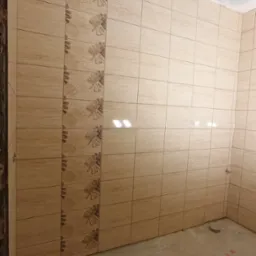 Ściany w łazience wyłożone beżowymi płytkami z motywem liści w pionowym pasie, surowy beton na podłodze, sufit w trakcie wykańczania.