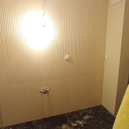 Bartłomiej Penk - Przygotowanie do remontu łazienki: ściany wyłożone pionowymi panelami, widoczne przyłącza wodne, podłoga w ciemnych, kwadratowych płytkach z efektem spękań, oświetlenie sufitowe.