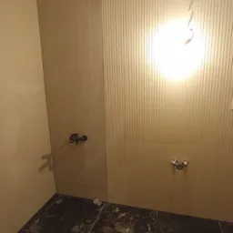 Ściany wyłożone beżowymi płytkami z fakturą pionowych pasków w łazience, czarna marmurkowa posadzka z płytek, widoczne przyłącza wodne i bateria.