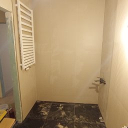 Bartłomiej Penk - Remont łazienki w trakcie: położone ciemne płytki podłogowe z marmurkowym wzorem, jasne płytki ścienne, grzejnik drabinkowy na ścianie, widoczne narzędzia i pozostałości materiałów budowlanych.