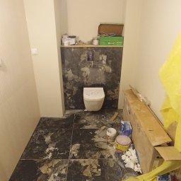 Bartłomiej Penk - Łazienka w trakcie prac: częściowo położone ciemne płytki imitujące marmur na podłodze i ścianie, wisząca toaleta, widoczne narzędzia i materiały budowlane, żółta folia ochronna.