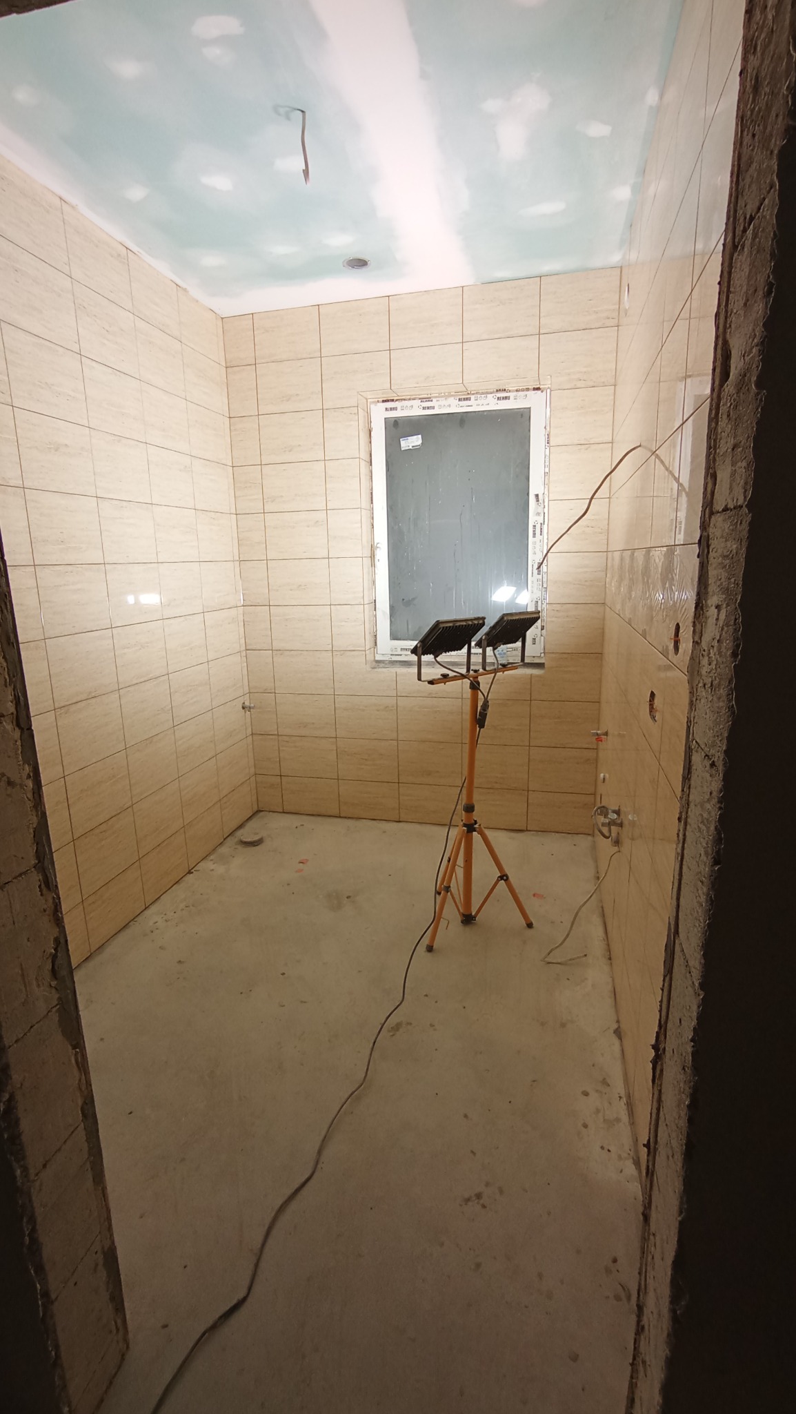 Niedokończona łazienka z beżowymi płytkami na ścianach, surowym sufitem z widocznymi łatami gipsu, okno w białej ramie i lampa budowlana na trójnogu.