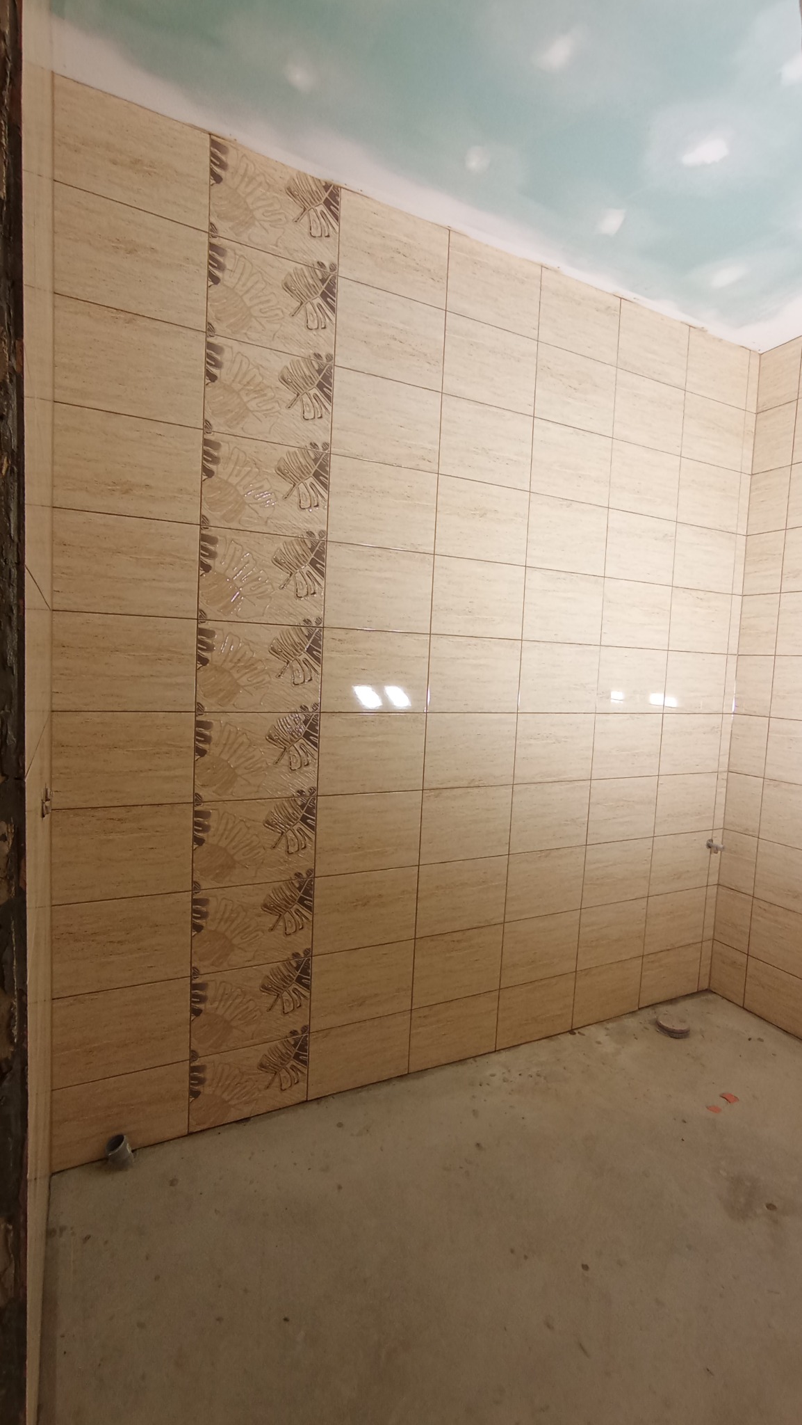 Ściany w łazience wyłożone beżowymi płytkami z motywem liści w pionowym pasie, surowy beton na podłodze, sufit w trakcie wykańczania.