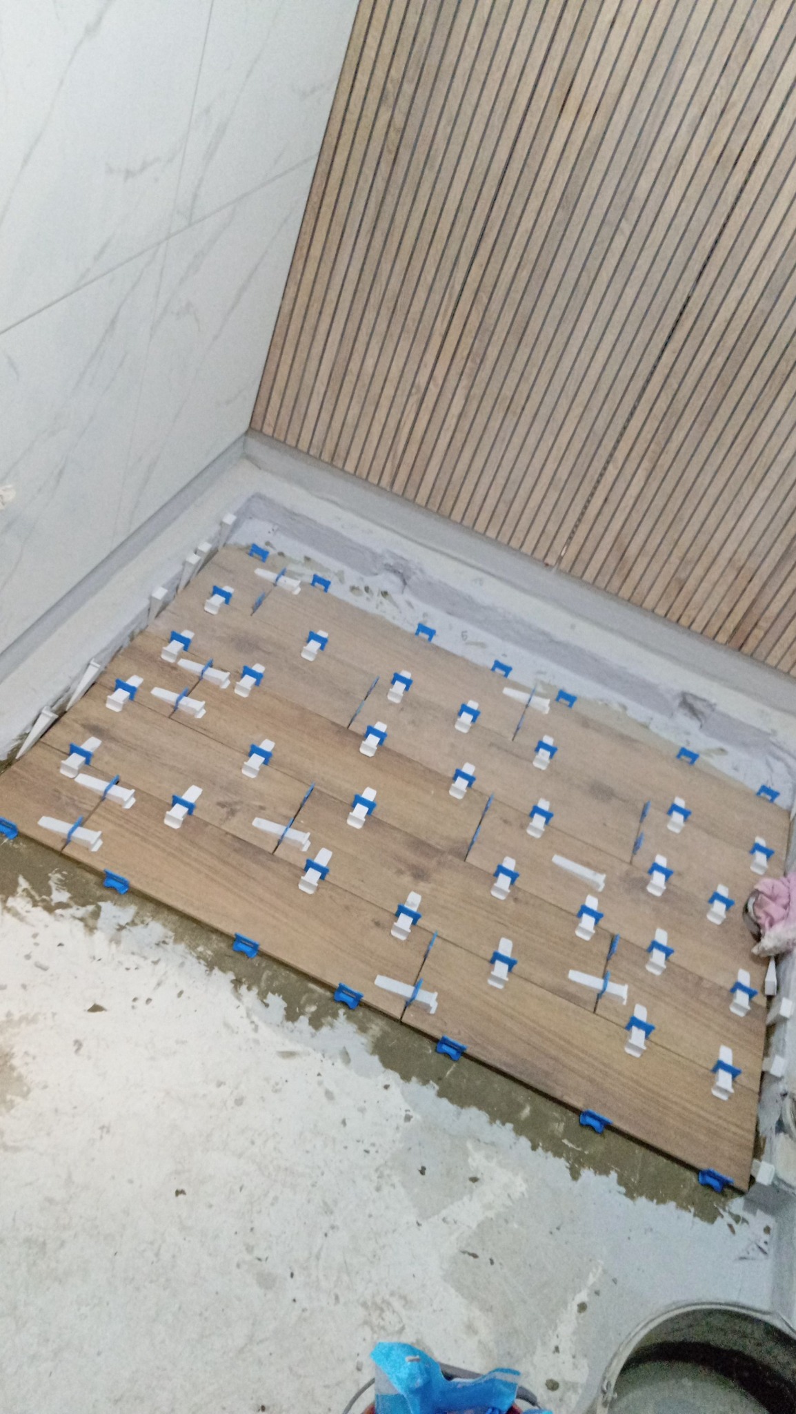 Układanie drewnopodobnych płytek podłogowych z użyciem niebieskich krzyżyków dystansowych, widoczne fragmenty ściany wyłożonej białym marmurem i drewnianą boazerią.