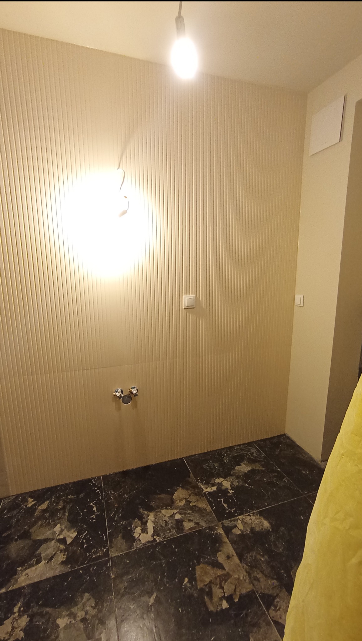 Przygotowanie do remontu łazienki: ściany wyłożone pionowymi panelami, widoczne przyłącza wodne, podłoga w ciemnych, kwadratowych płytkach z efektem spękań, oświetlenie sufitowe.