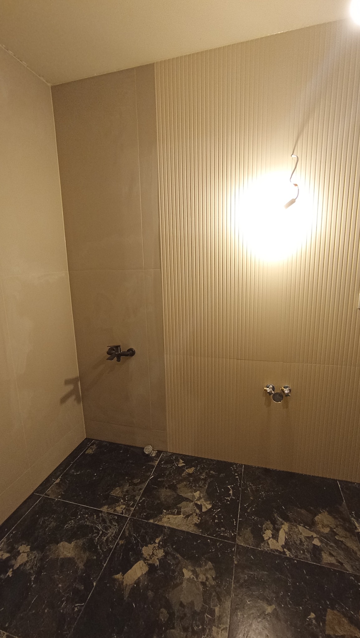 Ściany wyłożone beżowymi płytkami z fakturą pionowych pasków w łazience, czarna marmurkowa posadzka z płytek, widoczne przyłącza wodne i bateria.