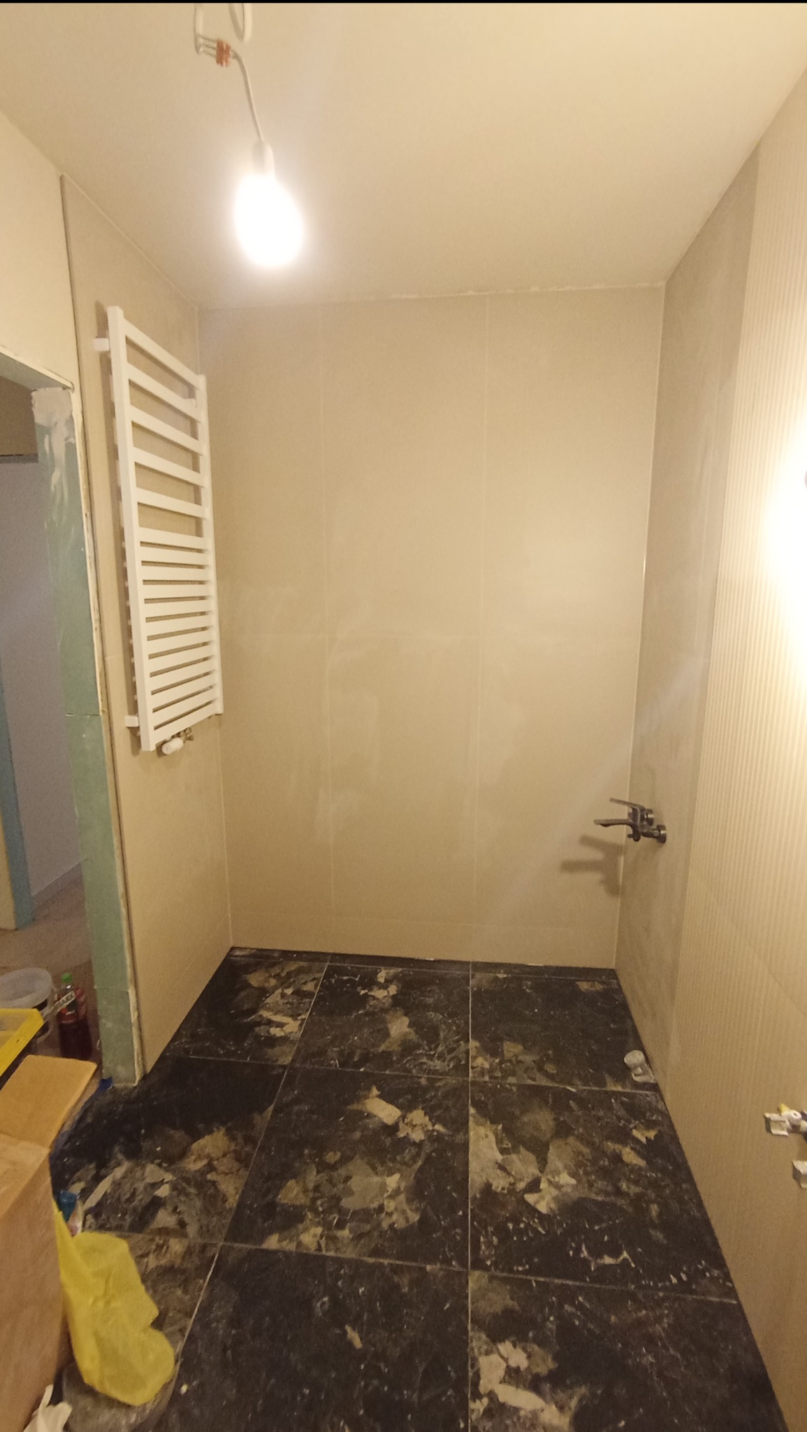 Remont łazienki w trakcie: położone ciemne płytki podłogowe z marmurkowym wzorem, jasne płytki ścienne, grzejnik drabinkowy na ścianie, widoczne narzędzia i pozostałości materiałów budowlanych.